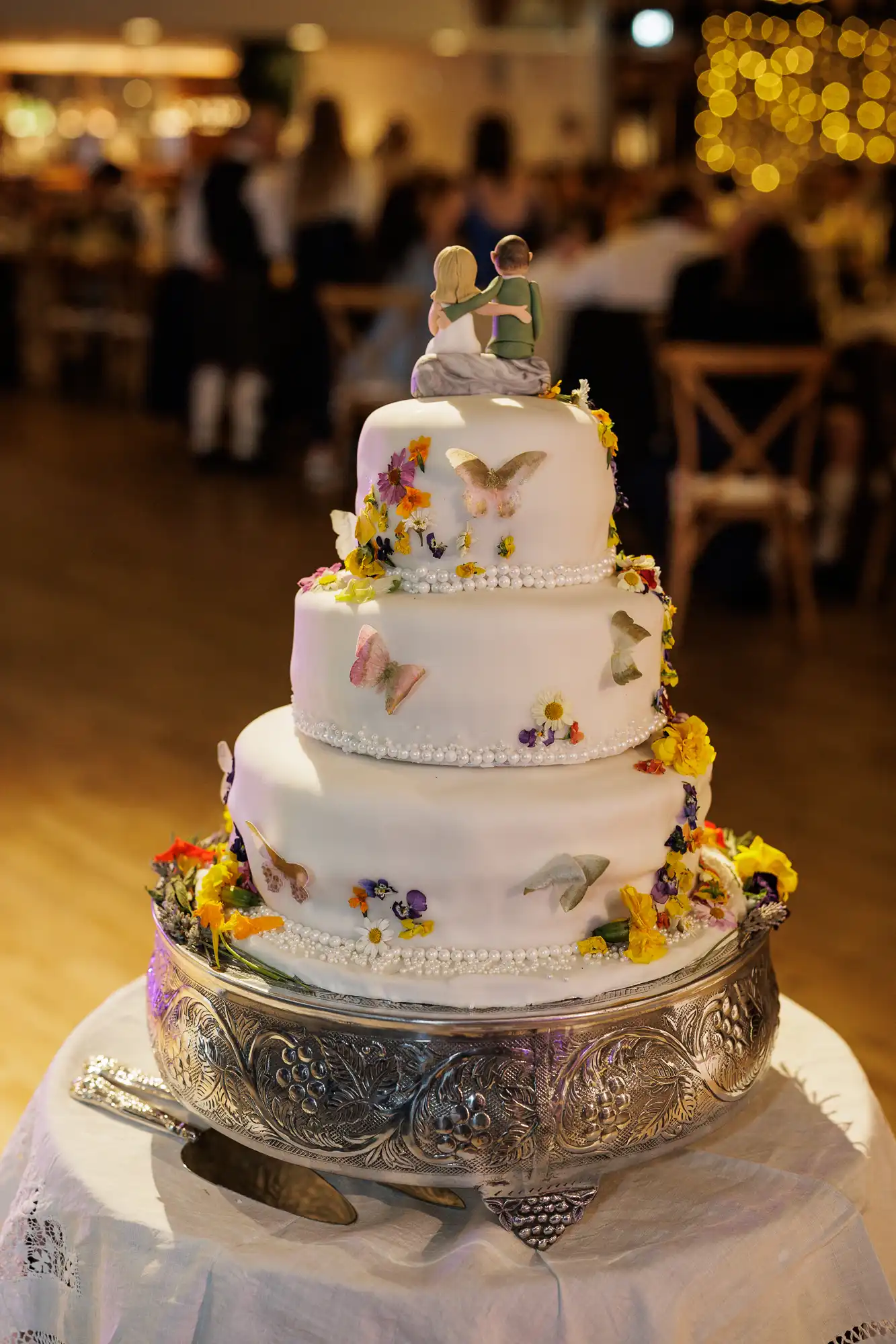 Three-tiered white wedding cake decorated with colourful edible flowers and butterfly designs, topped with a figurine of a bride and groom dancing. The cake sits on an ornate silver stand, with a cake knife on a white cloth-covered table. Blurred guests in background.