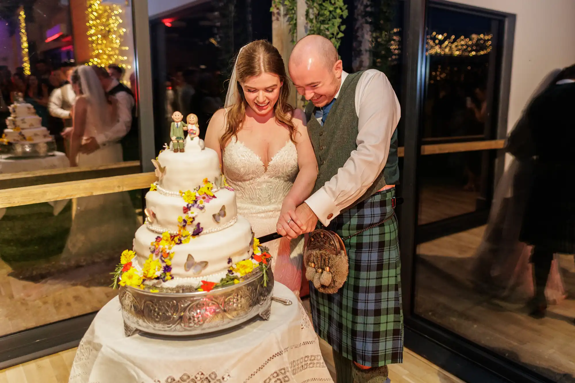 A smiling bride in a white lace gown and a groom in a green tartan kilt cut a white, three-tier wedding cake decorated with colourful flowers, standing on a silver tray. Guests and fairy lights are visible in the background.