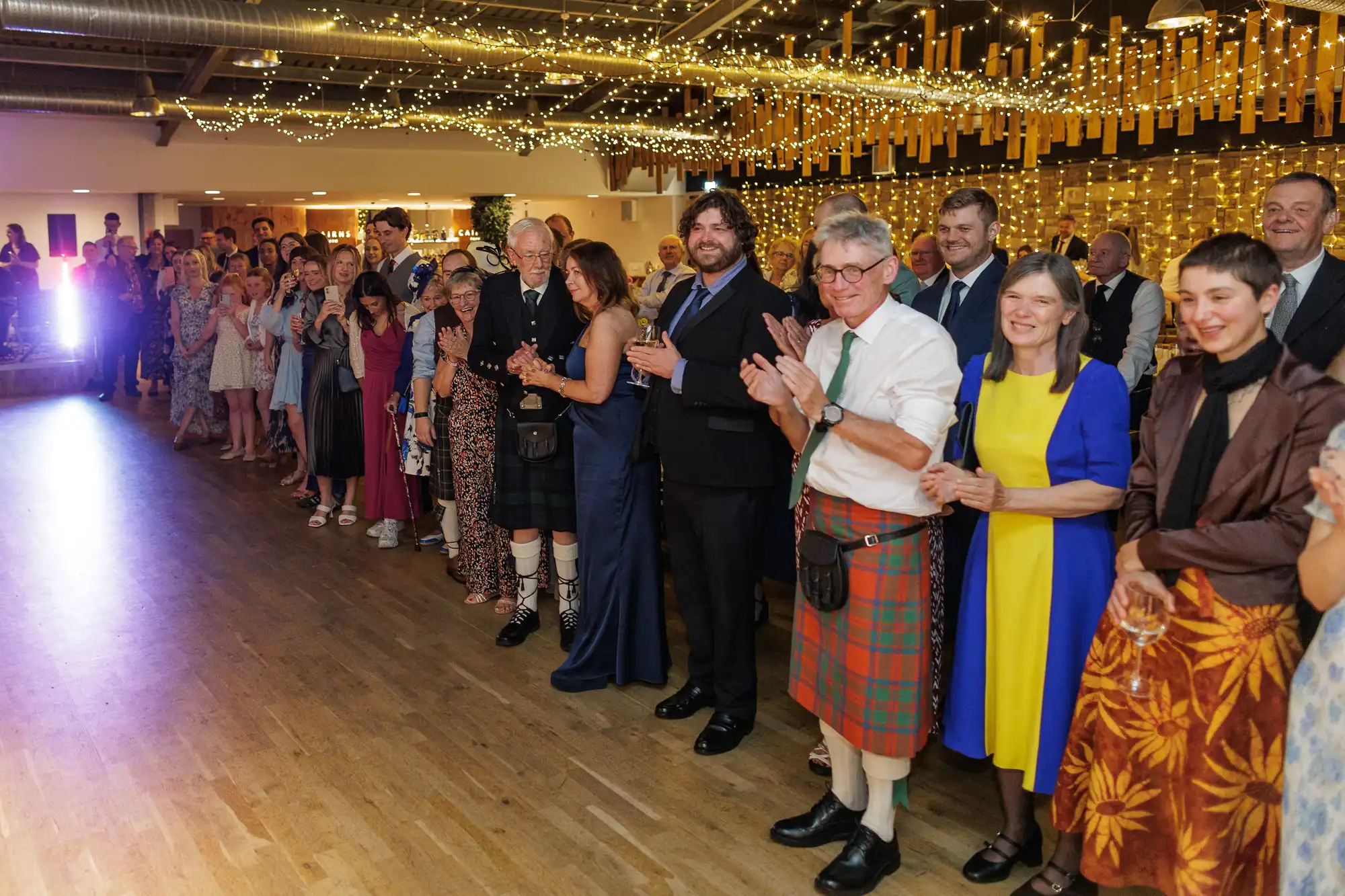 A large group of people dressed in formal and semi-formal attire, some in kilts, stand in a warmly lit hall decorated with fairy lights and hanging ribbons, smiling and applauding as they watch something off to the left.