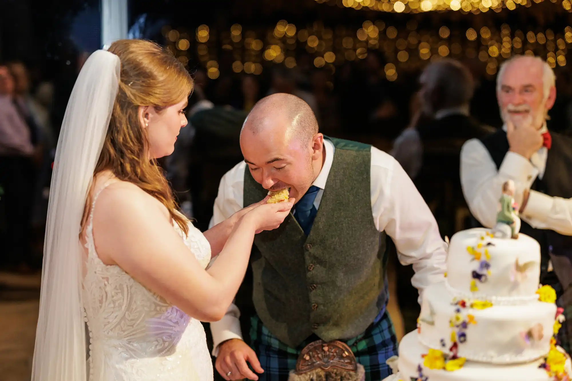 A bride in a white dress and veil feeds cake to a smiling groom in a waistcoat and kilt. He leans forward to take a bite. A decorated, floral wedding cake is beside them. Guests watch joyfully in the softly lit background.