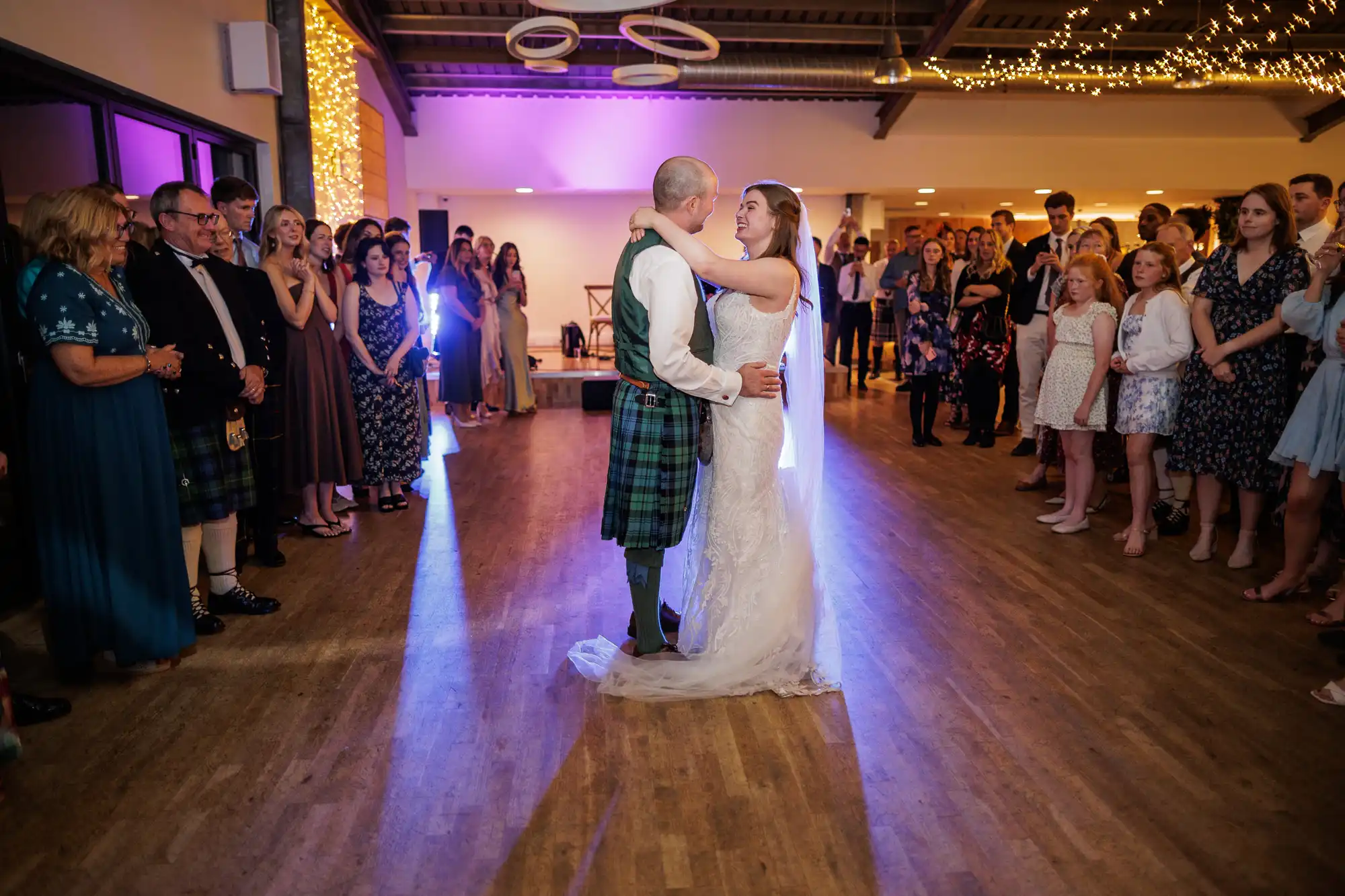 A bride and groom dance in the centre of a warmly lit room, surrounded by guests in formal attire. Fairy lights hang overhead, and people stand in a circle, smiling and watching the couple's first dance on a wooden floor.