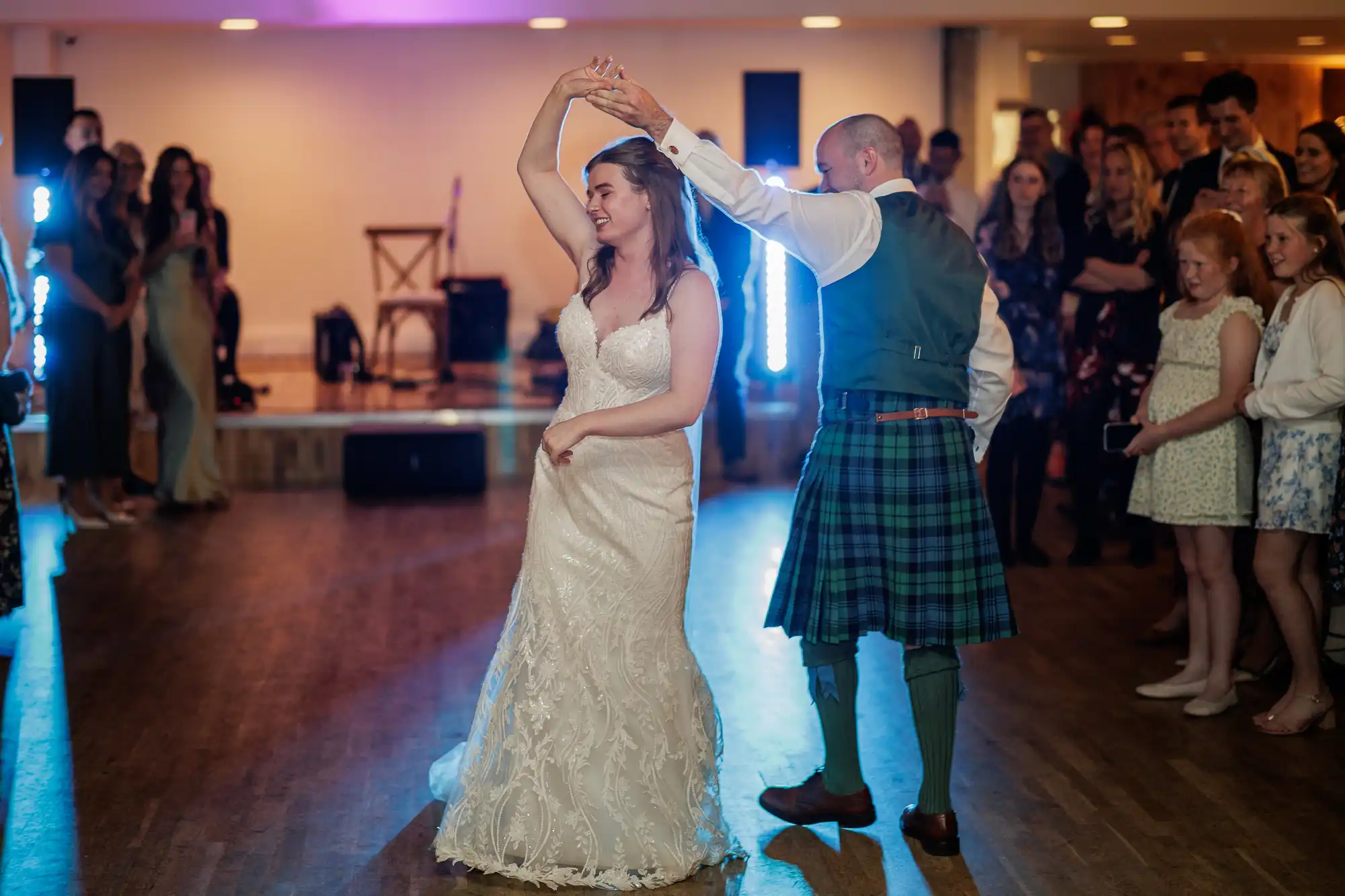 A bride in a white lace gown and a groom in a green tartan kilt dance together in a warmly lit room, surrounded by smiling guests. The groom twirls the bride, who looks happy. The wooden floor and stage with instruments are visible in the background.