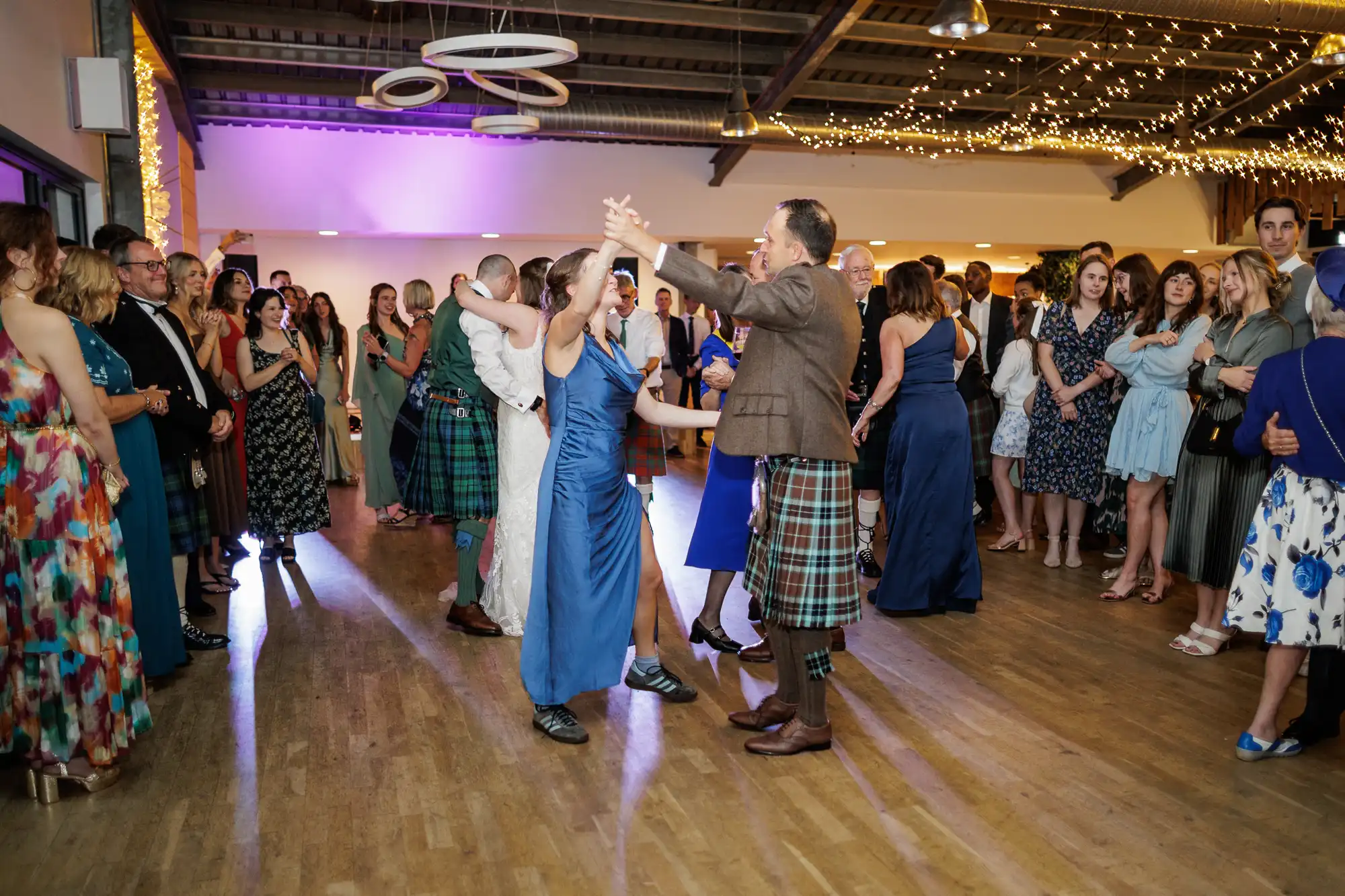 A lively group of people dance in pairs at an indoor event. Many men wear kilts, and women wear colourful dresses. Guests stand around watching, fairy lights hang from the ceiling, and the atmosphere is festive and joyful.