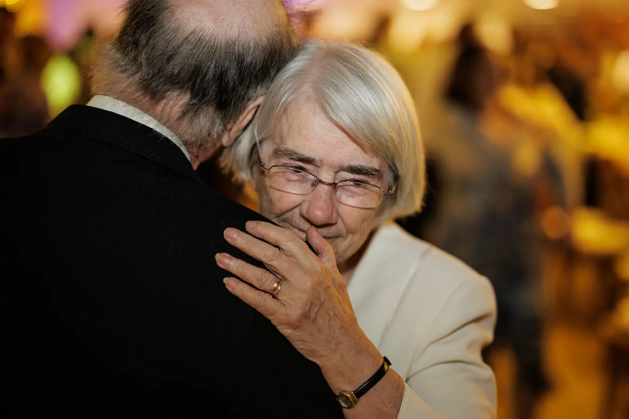 An elderly woman with glasses and white hair hugs a man with thinning hair, wearing a dark suit. She appears emotional, with a slight smile and teary eyes. The background is softly blurred, suggesting a warm, intimate gathering or event.