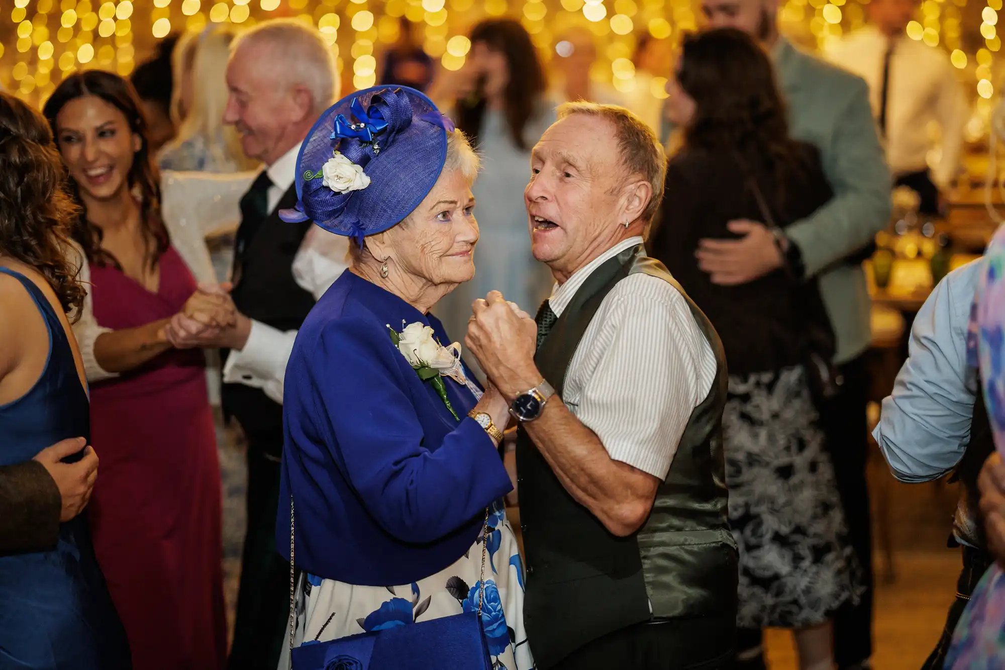 An elderly couple dressed formally dance together at a lively, warmly lit celebration. The woman wears a blue floral dress and matching hat with flowers; the man wears a waistcoat and white shirt. Smiling guests dance in the blurred, festive background with fairy lights.