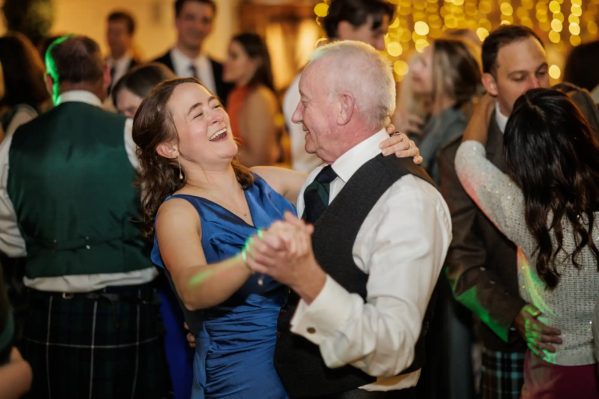 A young woman in a blue dress and an older man in a white shirt and waistcoat joyfully dance together at a lively event. Smiling guests surround them, and warm, golden fairy lights glow in the softly blurred background.