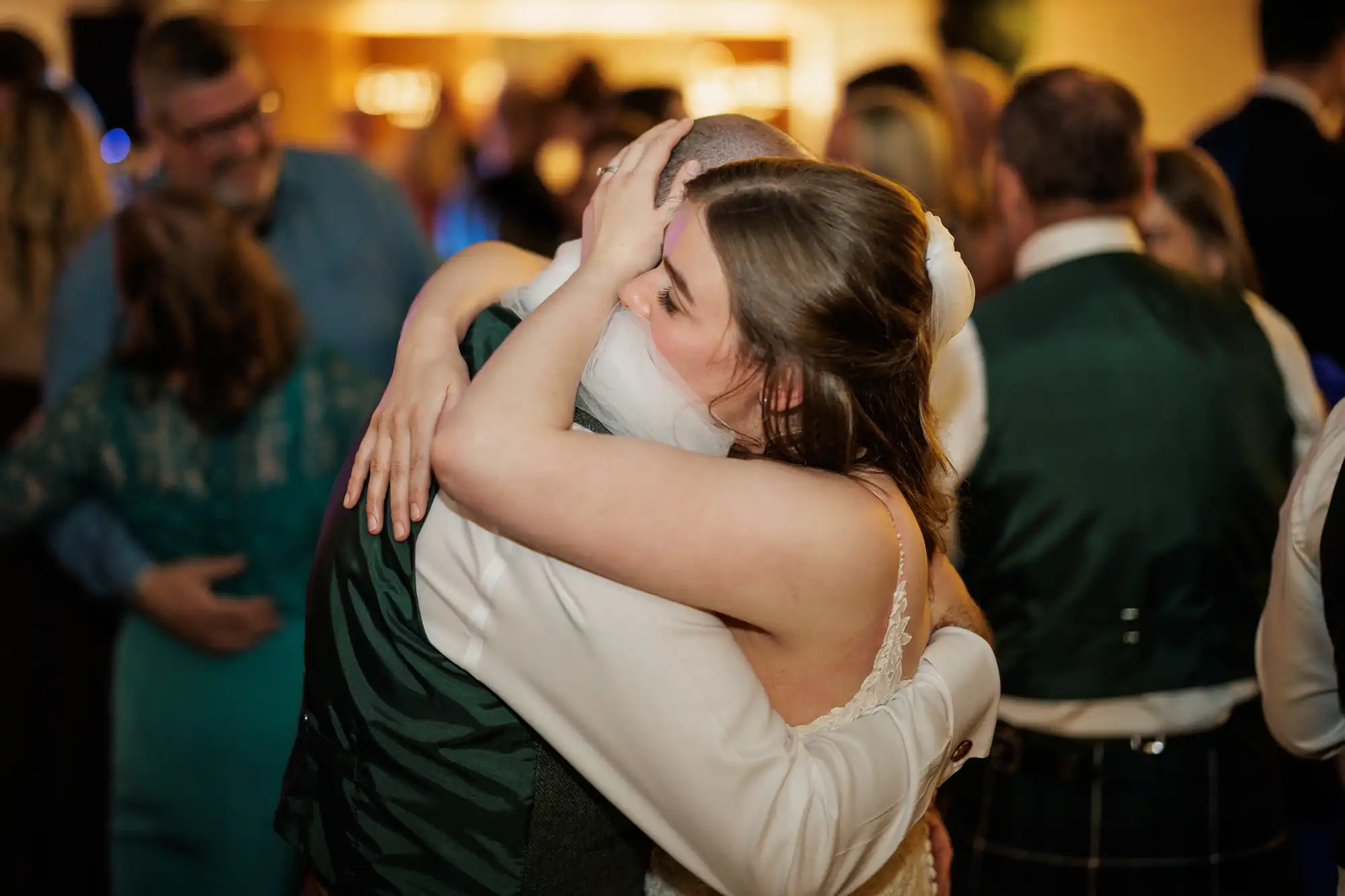 A bride in a white dress hugs a man in a dark waistcoat tightly at a crowded indoor event. Her eyes are closed and her hand is on his head, conveying deep emotion. Other guests, slightly out of focus, mingle in the warmly lit background.