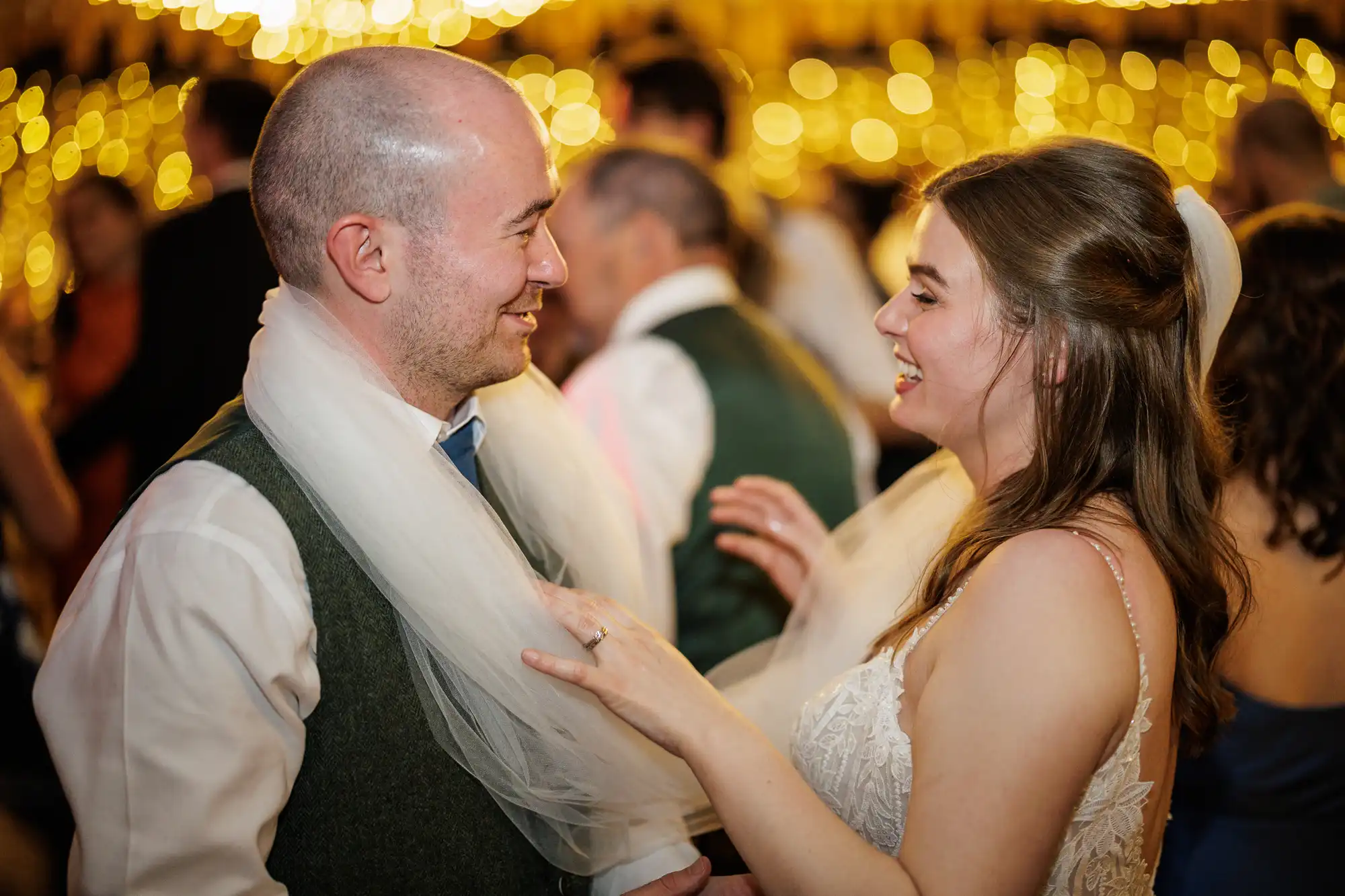 A smiling bride and groom face each other at their wedding reception, laughing as the bride’s veil is draped playfully around their shoulders. Soft golden lights blur in the background, and other guests are visible, celebrating together.