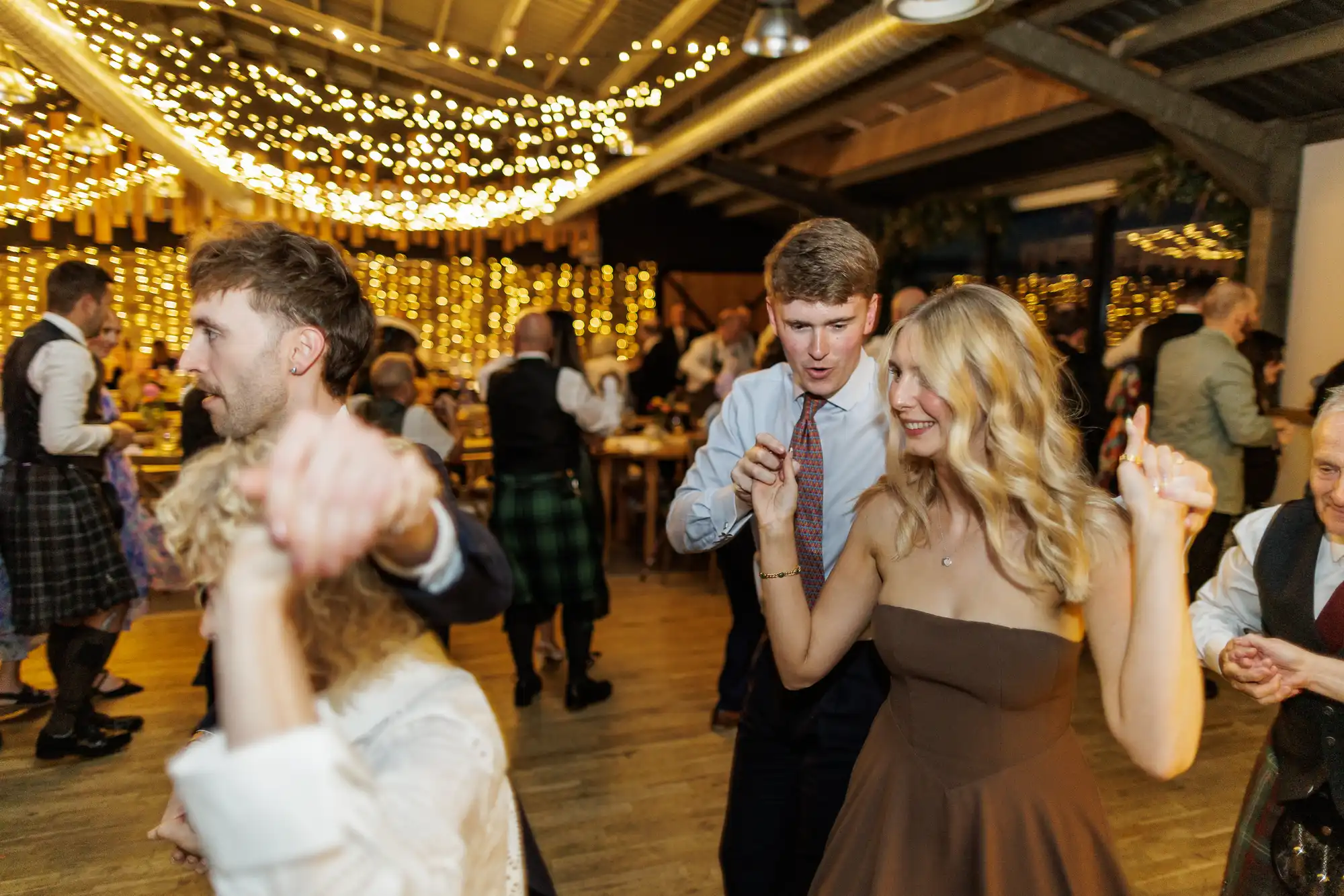 People dance joyfully at a festive indoor event with fairy lights and golden decorations. The atmosphere is lively, with couples and groups on the wooden floor. A smiling woman in a brown dress and a man in a blue shirt are in the foreground, dancing together.