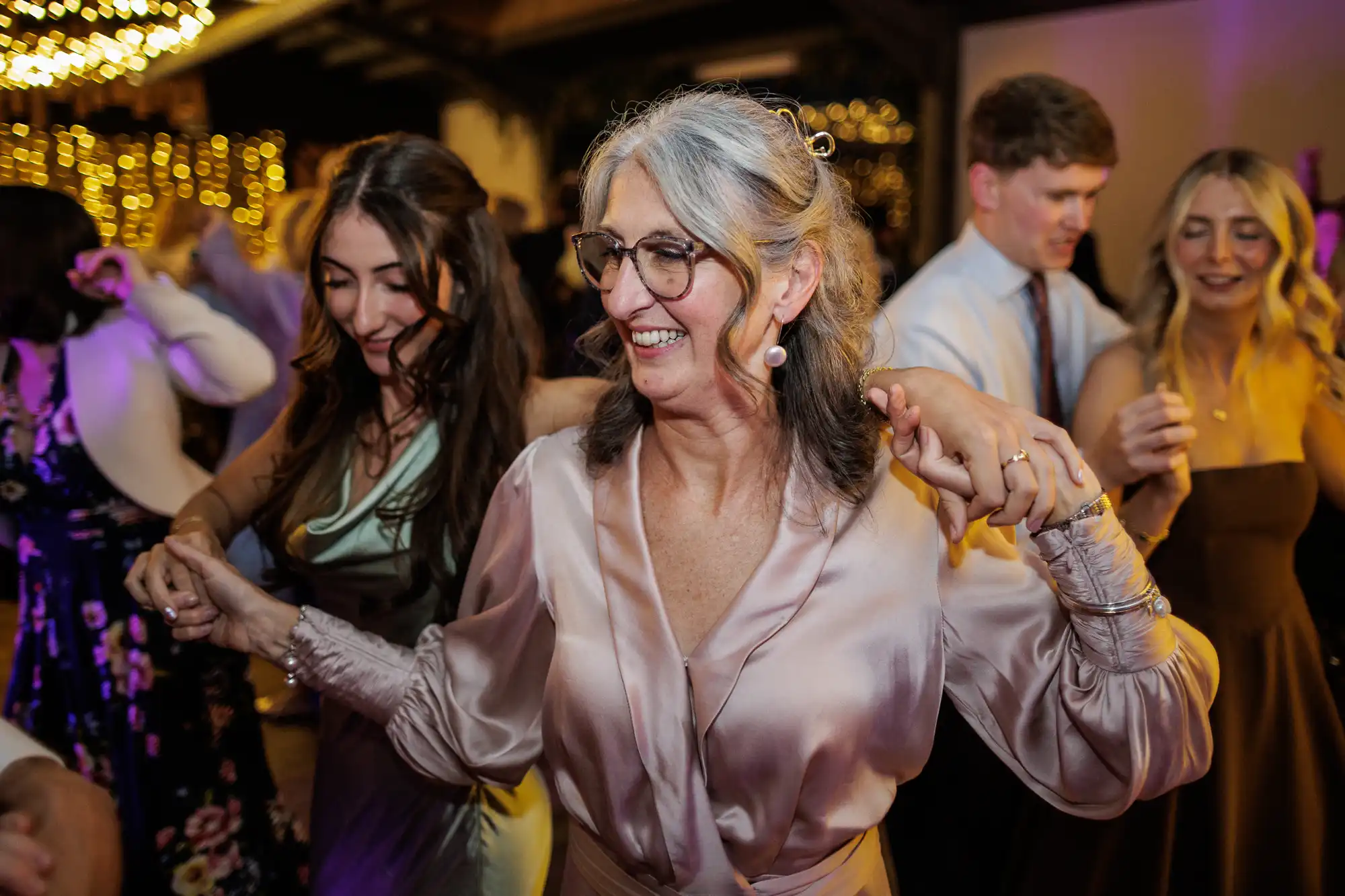 A joyful older woman with grey hair and glasses, wearing a silky pink blouse, dances and holds hands with two younger women at a lively indoor event. Warm lights and other smiling people are visible in the festive background.