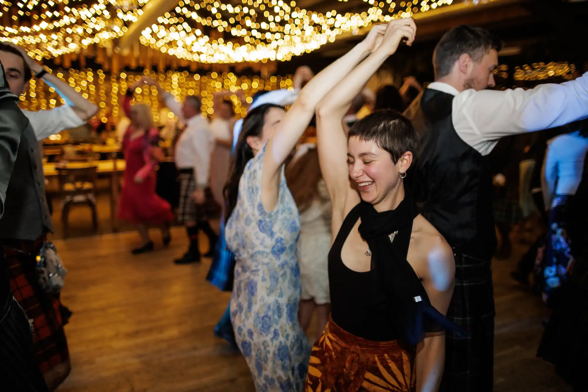 People dance joyfully under warm, twinkling fairy lights at a lively indoor event. Two women spin each other, smiling, surrounded by others in festive attire, including kilts and dresses, on a wooden floor. The atmosphere is vibrant and celebratory.
