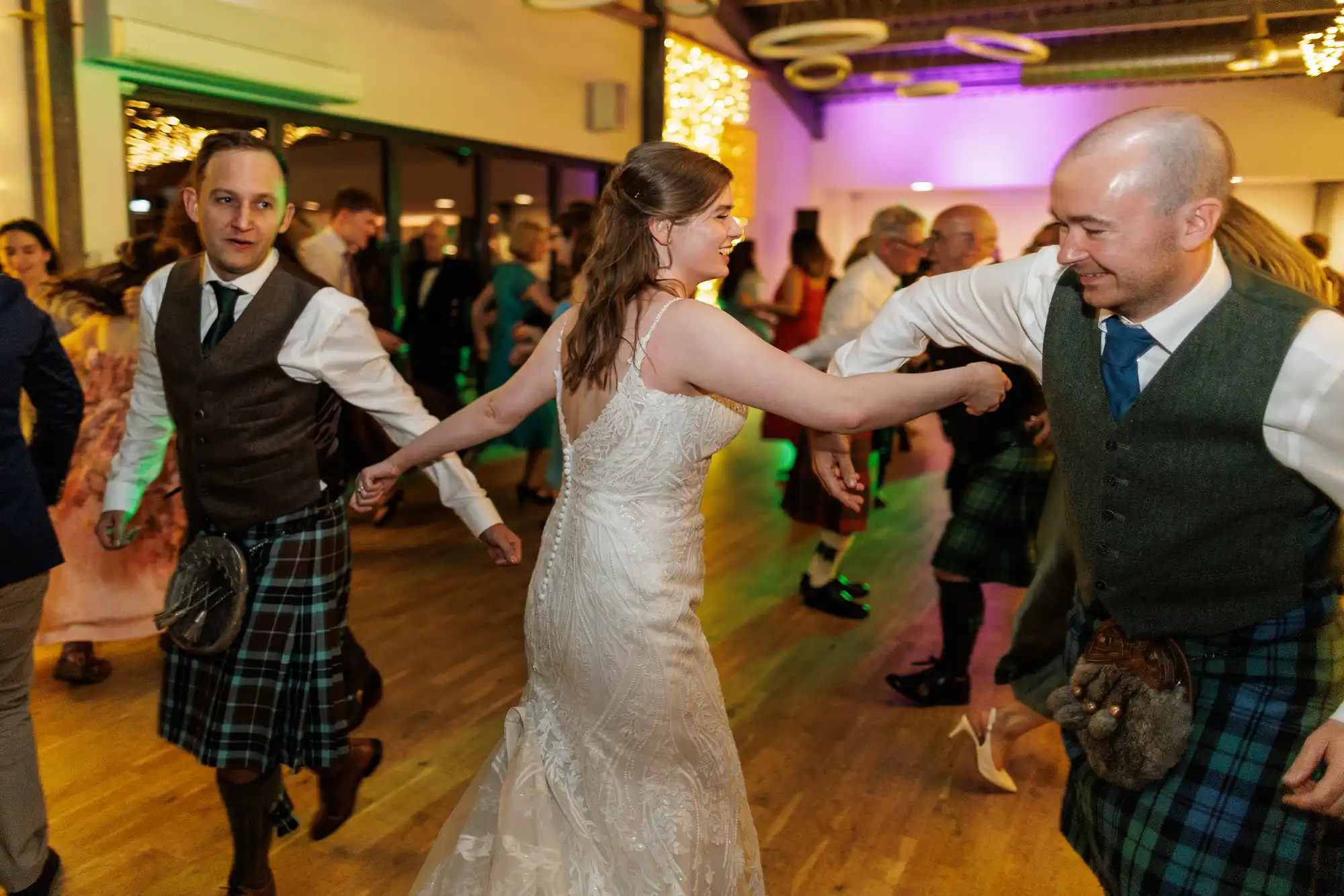 A bride in a white dress dances with two men in kilts and waistcoats at a lively indoor wedding reception, surrounded by other guests on a wooden floor, colourful lights illuminating the scene.