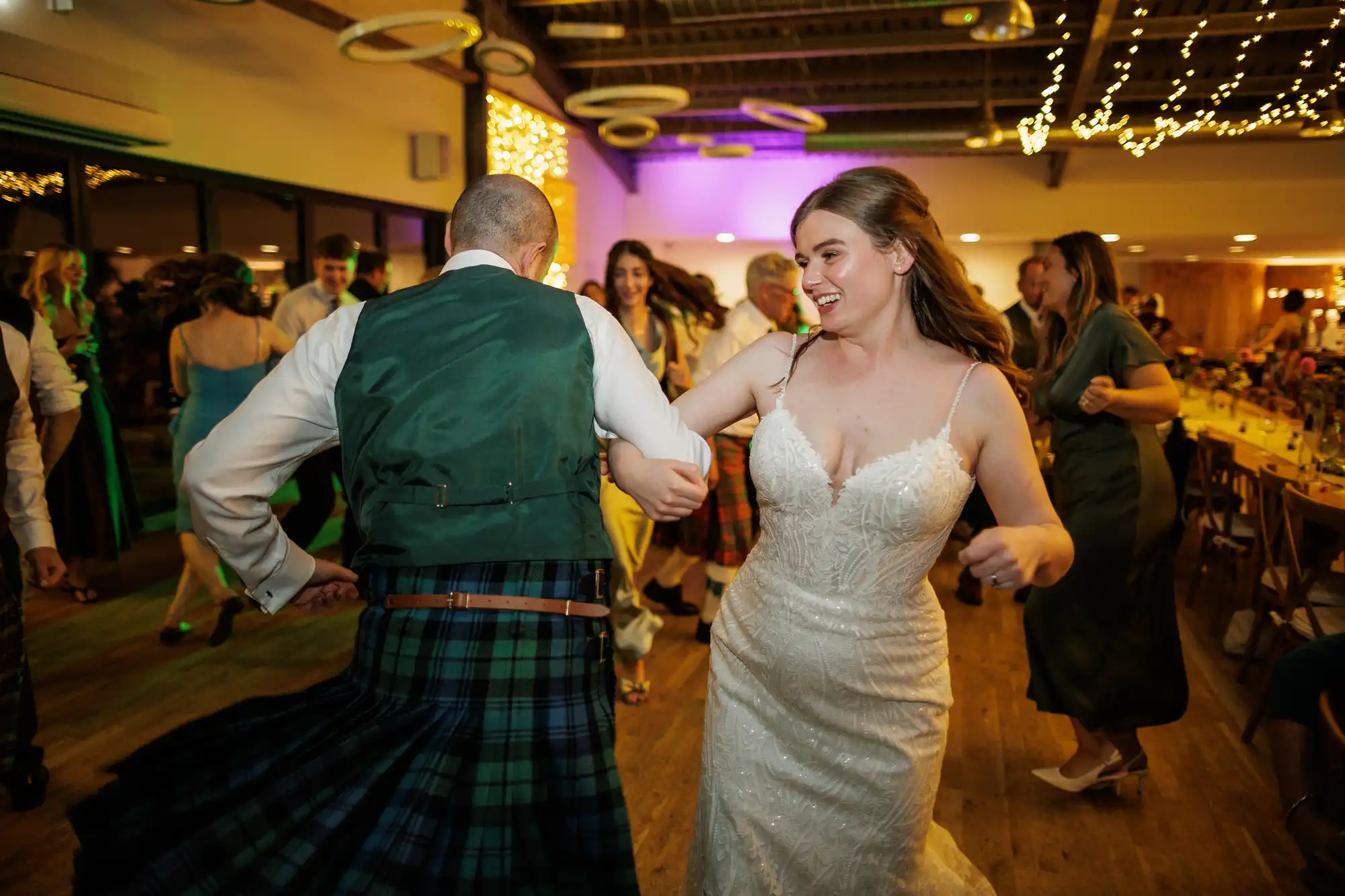 A bride in a white dress and a man in a green waistcoat and tartan kilt joyfully dance together at a lively wedding reception, surrounded by guests under fairy lights in a warmly lit venue.