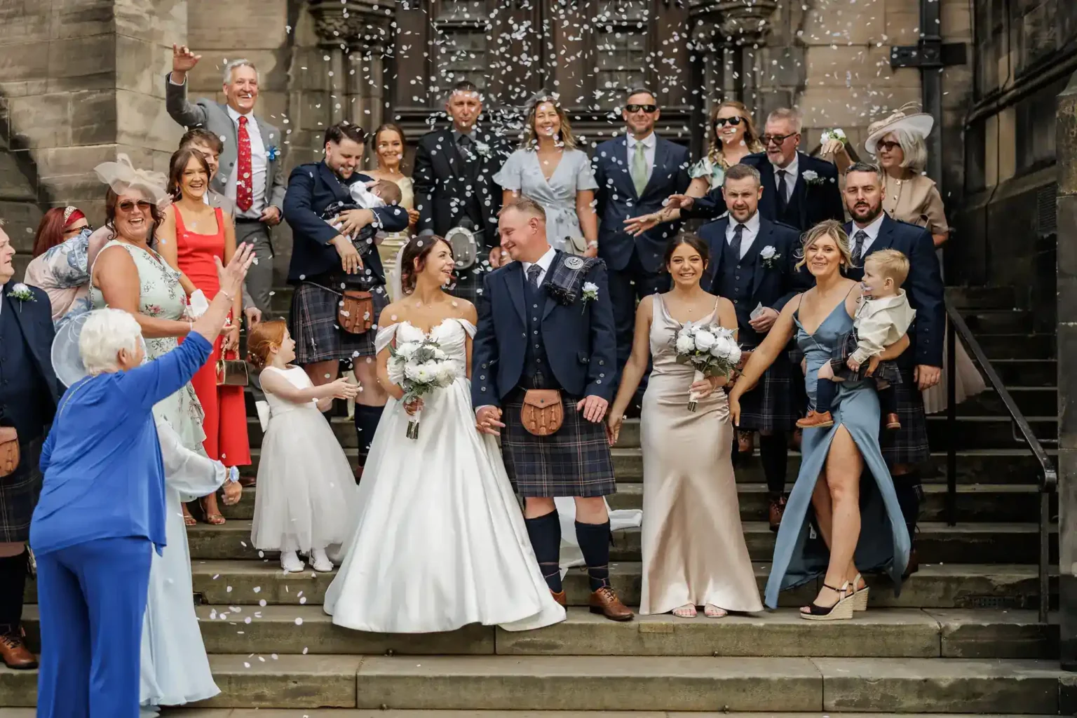 A wedding party poses on stone steps, with the bride and groom at the centre. Bridesmaids in pale dresses and men in kilts surround them. Guests smile, some throwing confetti. Two small children and an older woman in blue are visible. The mood is joyful and celebratory.