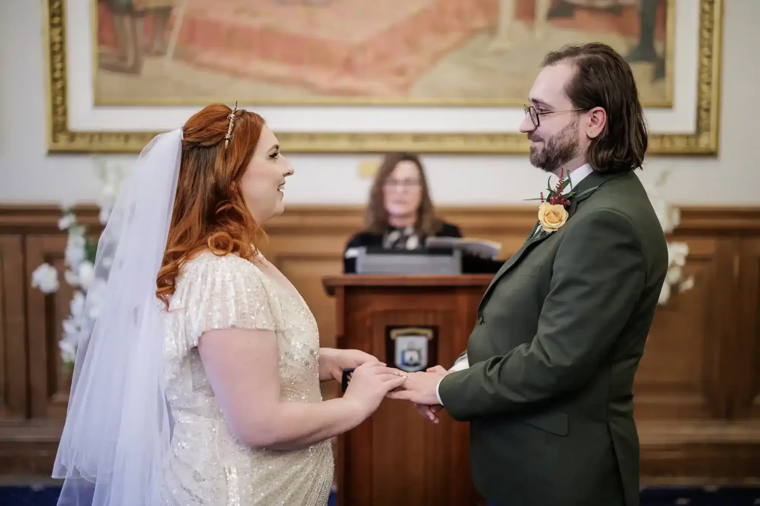 A bride in a white, beaded gown and veil holds hands with a groom in a green suit and buttonhole. They face each other, smiling, with a woman officiating in the background. Elegant flowers and a framed painting decorate the room.