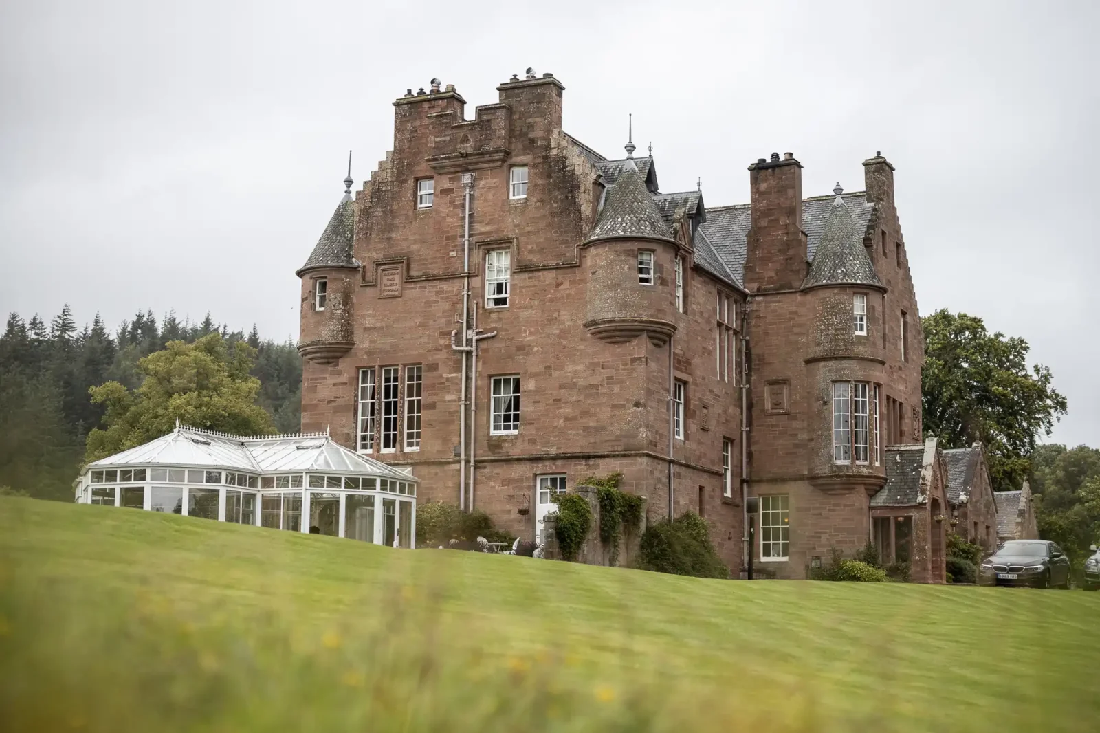 A large, historic stone mansion with turrets and pitched roofs sits on a green lawn. Attached to the left side is a glass conservatory. The sky is overcast, and trees are visible in the background.