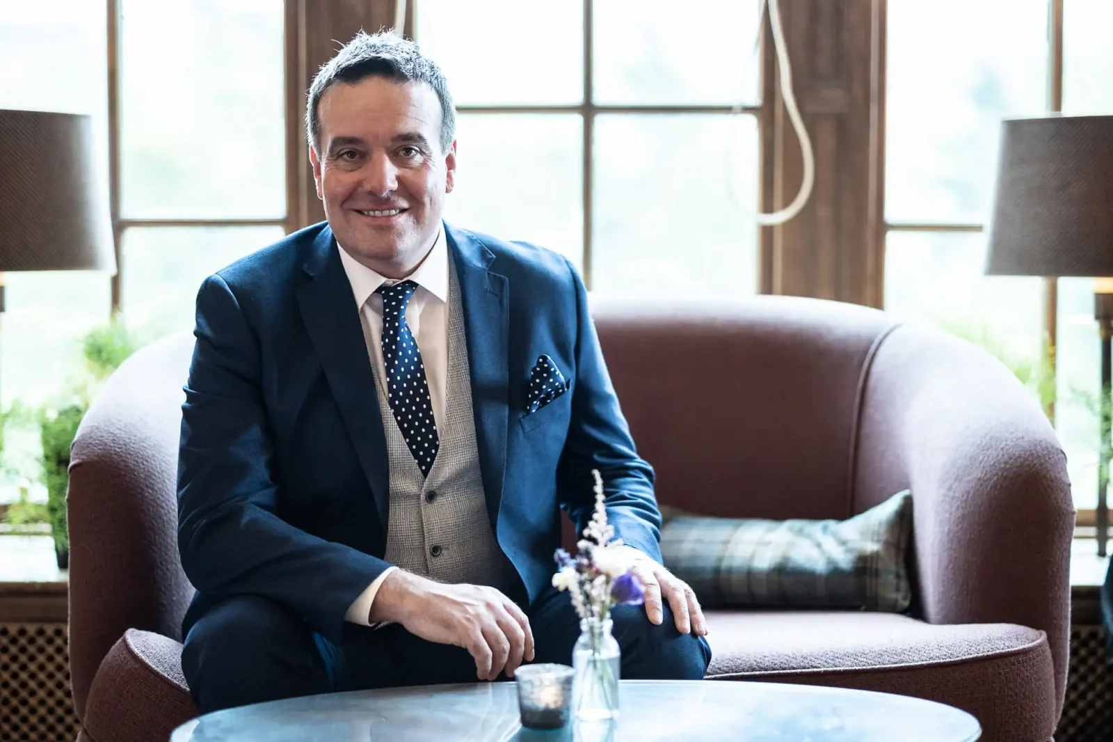 A man in a navy suit with a spotty tie sits on a mauve sofa, smiling at the camera. Behind him are large windows with natural light, two lamps, and a small round table in front with a candle and a small flower arrangement.