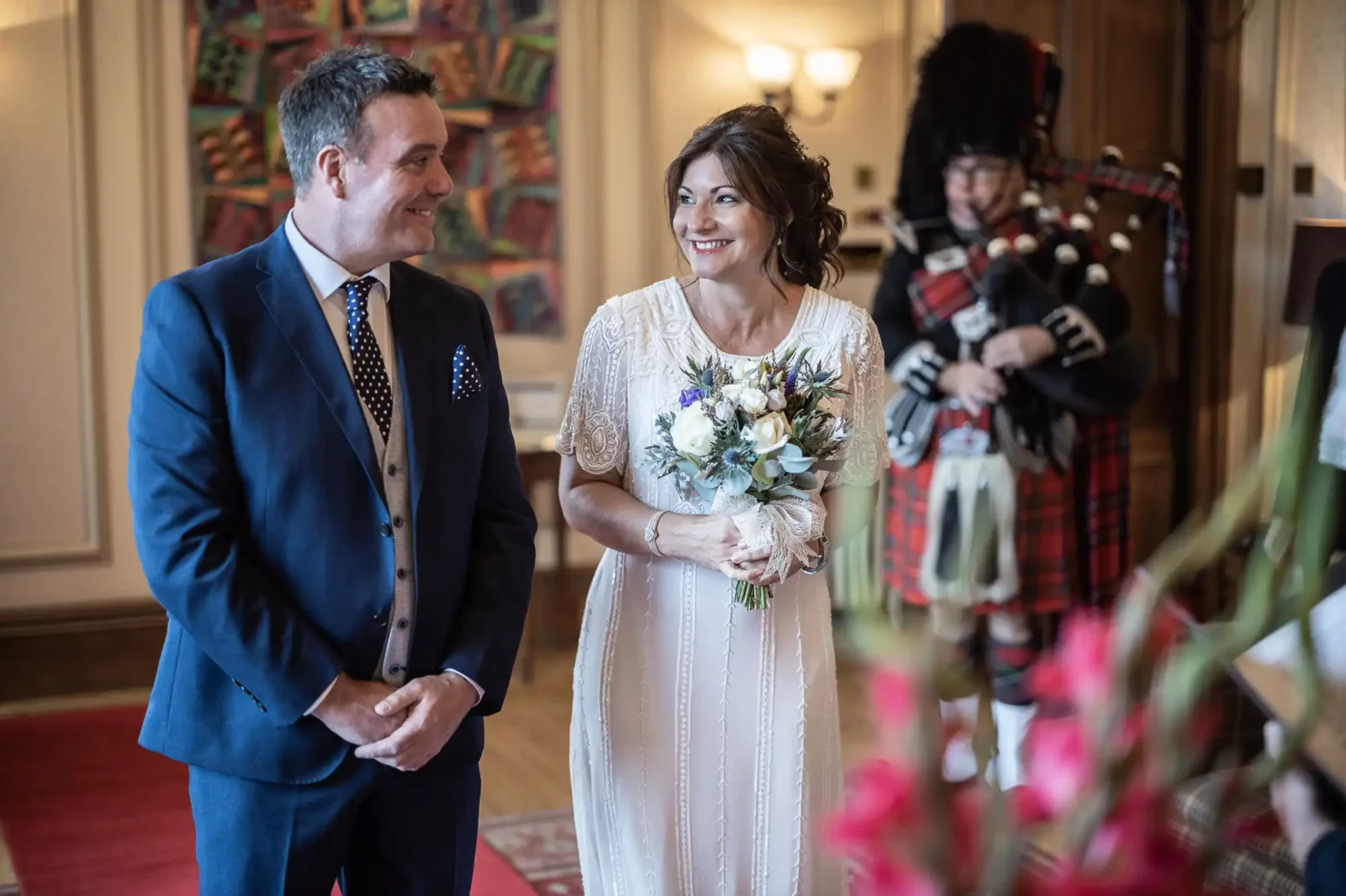 A smiling bride in a white dress holding a bouquet stands beside a groom in a navy suit and spotted tie. Both look at each other warmly. In the blurred background, a bagpiper in traditional Scottish attire plays. The setting is elegant and warmly lit.