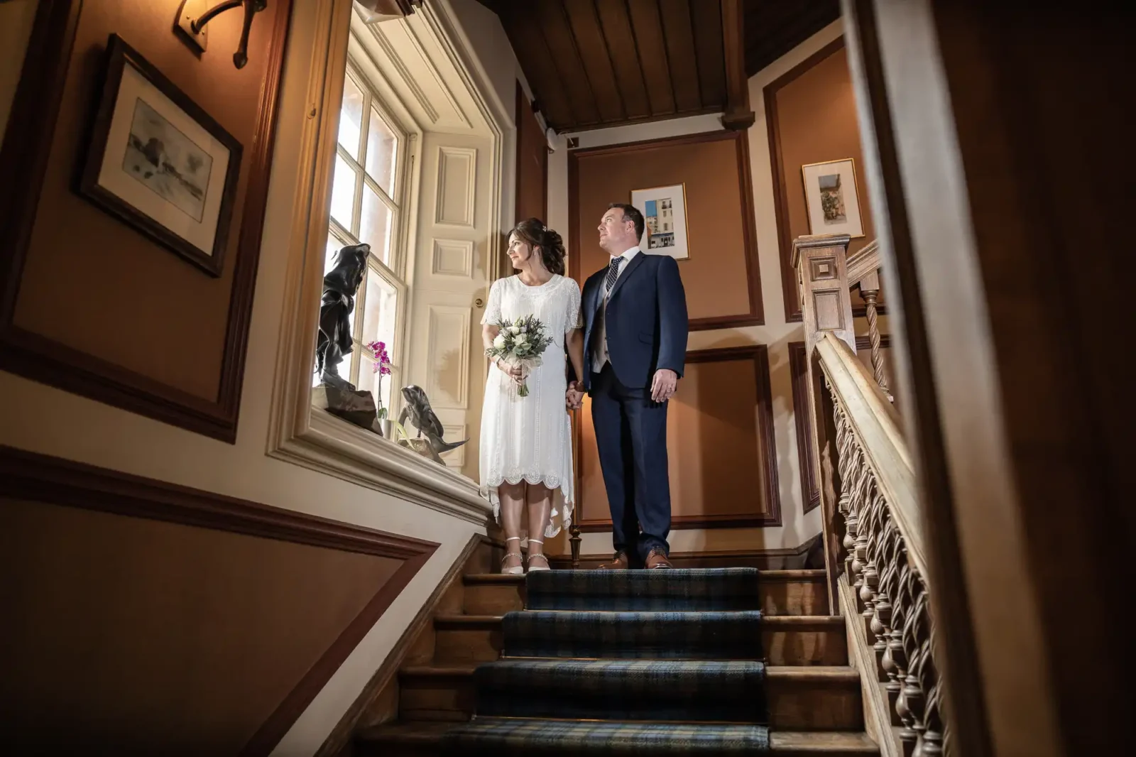 A bride in a white dress holding a bouquet and a groom in a navy suit stand side by side on a wooden staircase, gazing out of a sunlit window. The staircase has patterned carpet and ornate bannisters; framed art hangs on the brown-panelled walls.