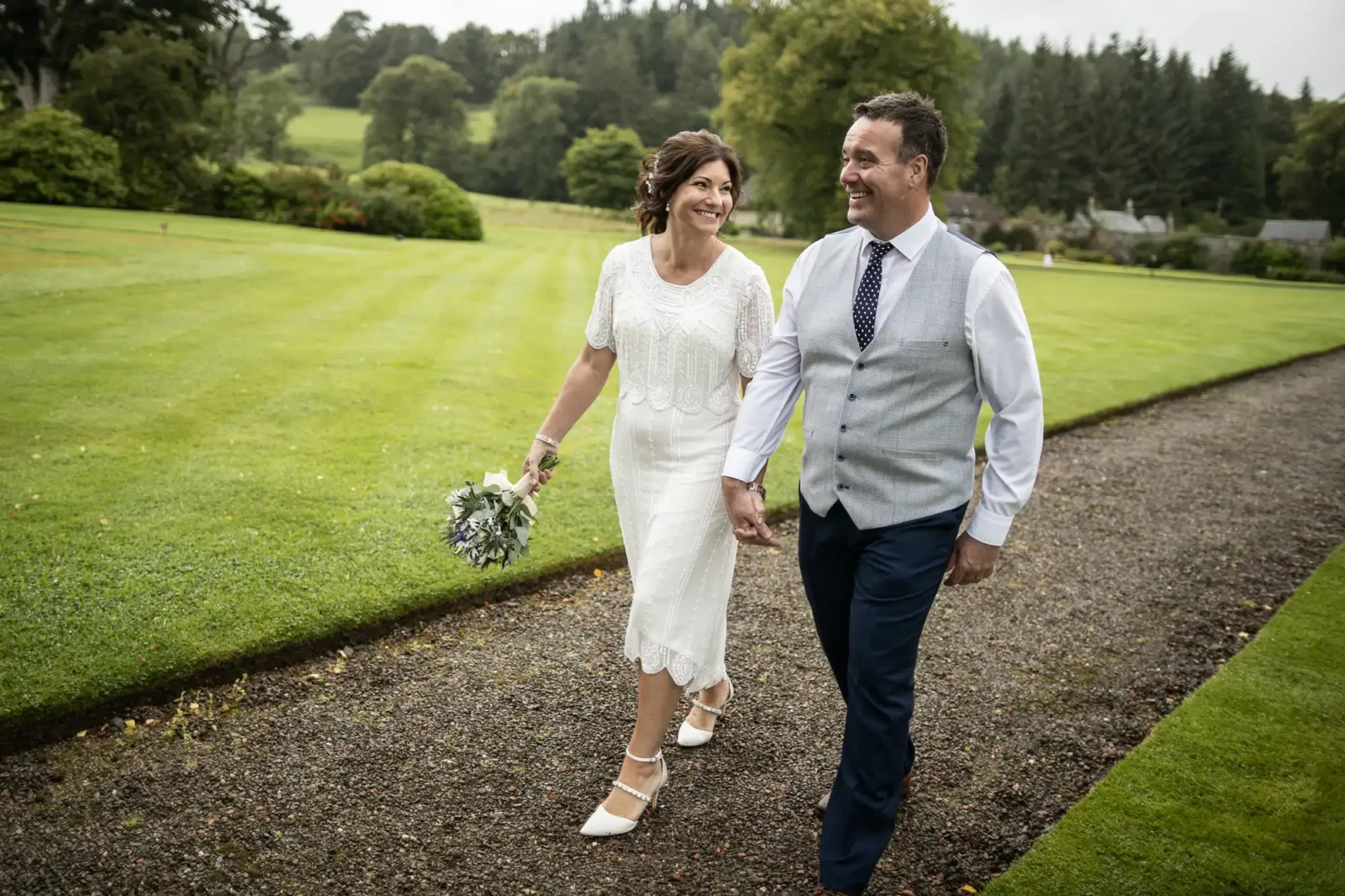 A smiling bride in a white dress holds a bouquet and walks hand-in-hand with a groom in a waistcoat, tie, and dark trousers along a gravel path in a green garden, with trees and grass in the background. Both look happy and relaxed.