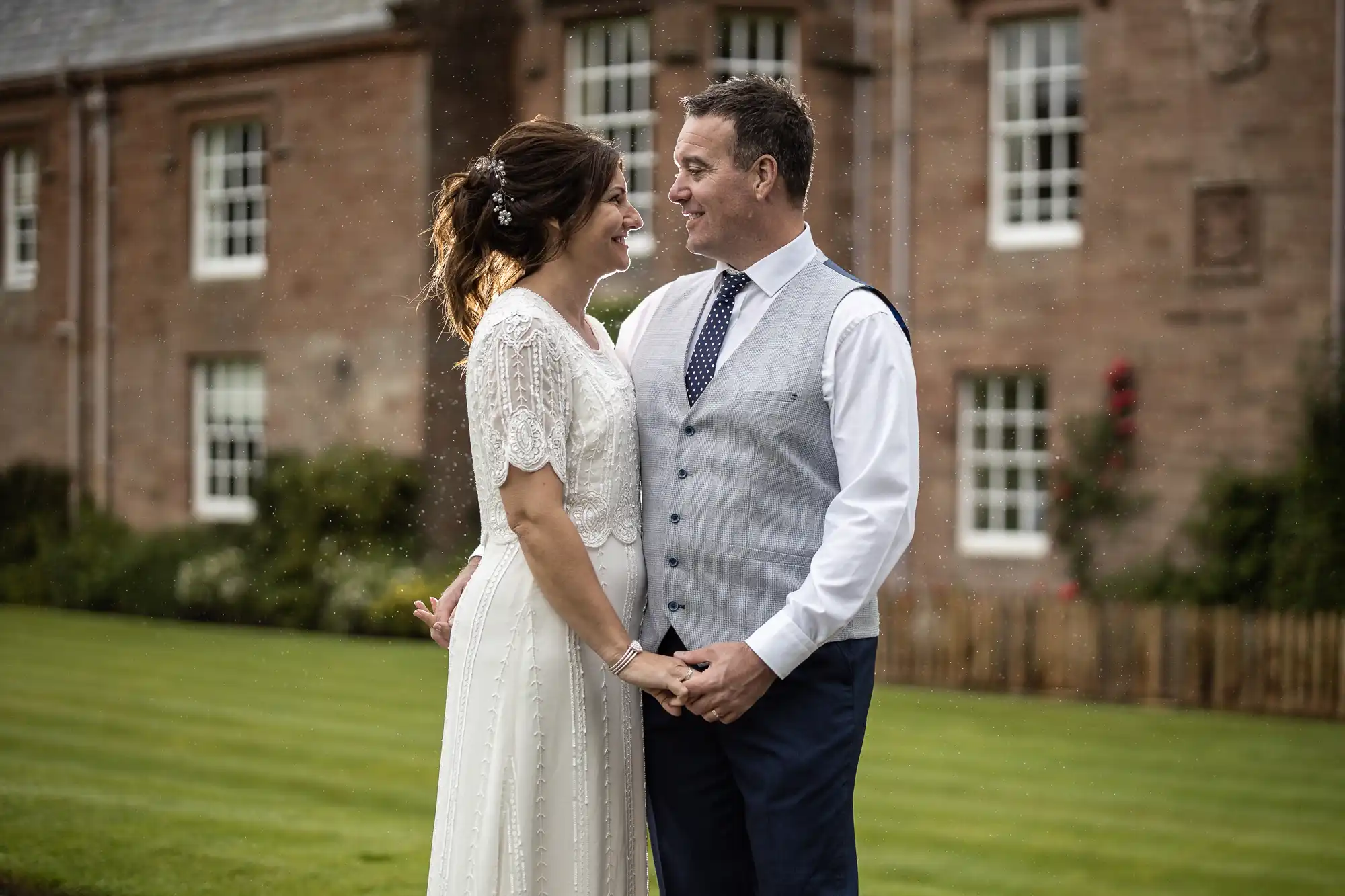 Cringletie wedding photographer image of a bride and groom stand outdoors holding hands, smiling and gazing at each other. The bride wears a white dress with lace details; the groom wears a white shirt, tie, and light grey waistcoat. Behind them is a brick building, green lawn, and some rain falling.