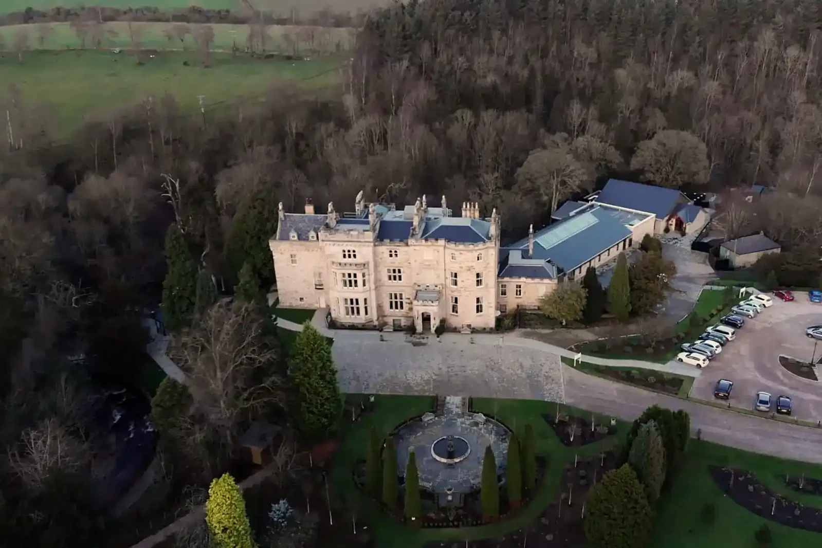 Aerial view of a large stone mansion with multiple chimneys, a circular drive, manicured gardens, and a central fountain, surrounded by trees and fields, with parked cars visible to one side.