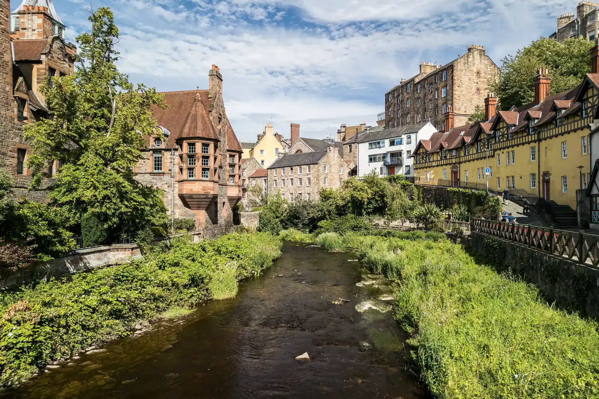 A clear stream flows through a lush green area, bordered by historic stone and yellow buildings. Trees and bushes line the banks, and rooftops and old architecture rise against a partly cloudy blue sky.