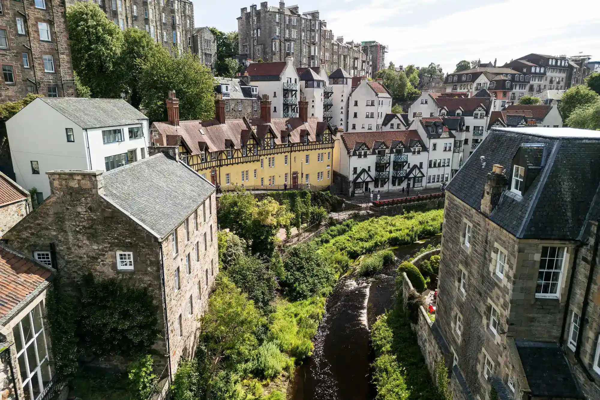 A small river winds through a lush, green area surrounded by historic stone buildings and colourful houses with steep roofs in a scenic European town on a sunny day.