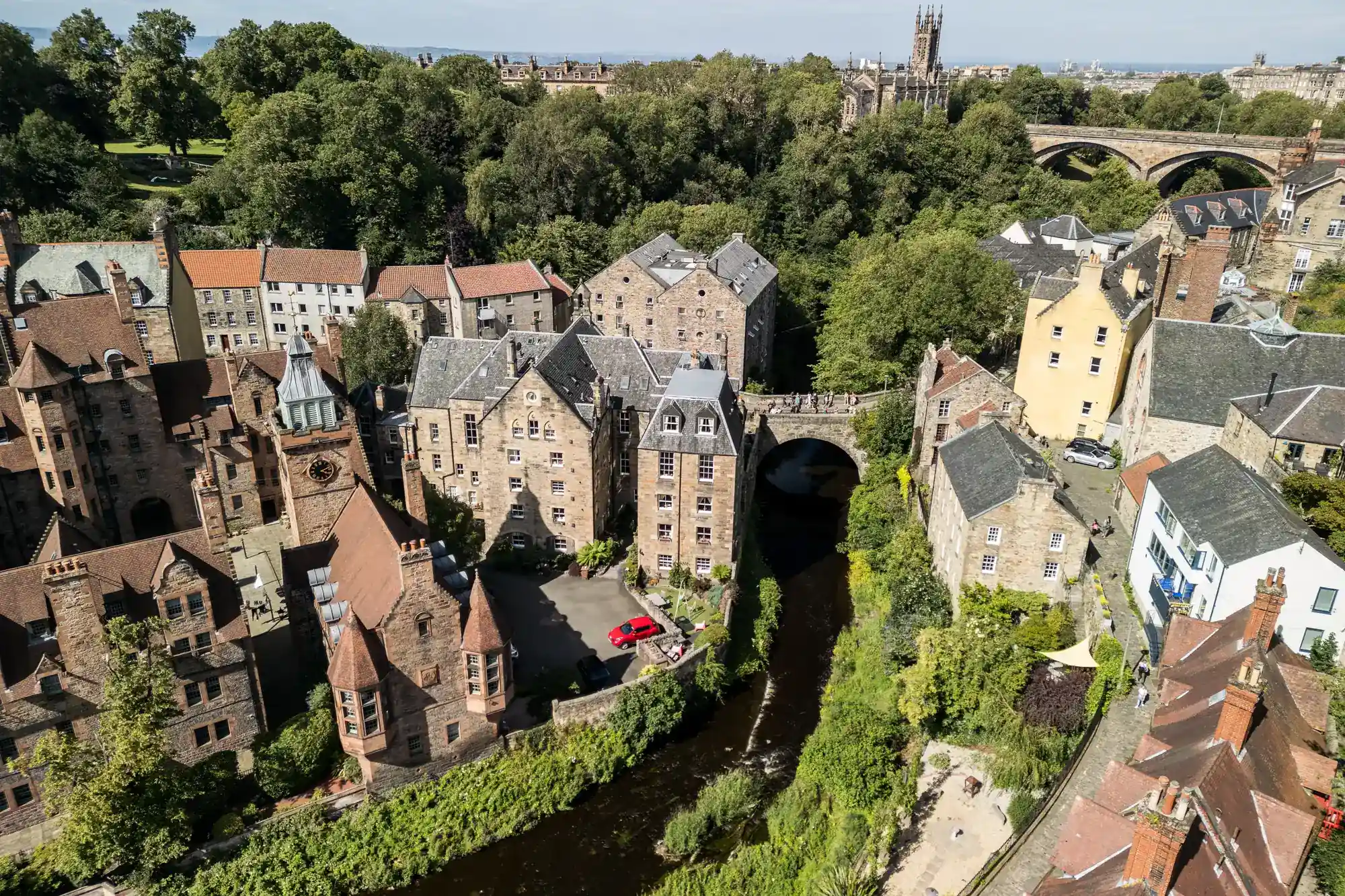 Aerial view of a historic stone village with pointed roofs, a river flowing through, lush trees, and a stone bridge in the background. A red car is parked near the river. The scene is bright and peaceful, showing a mix of greenery and old architecture.