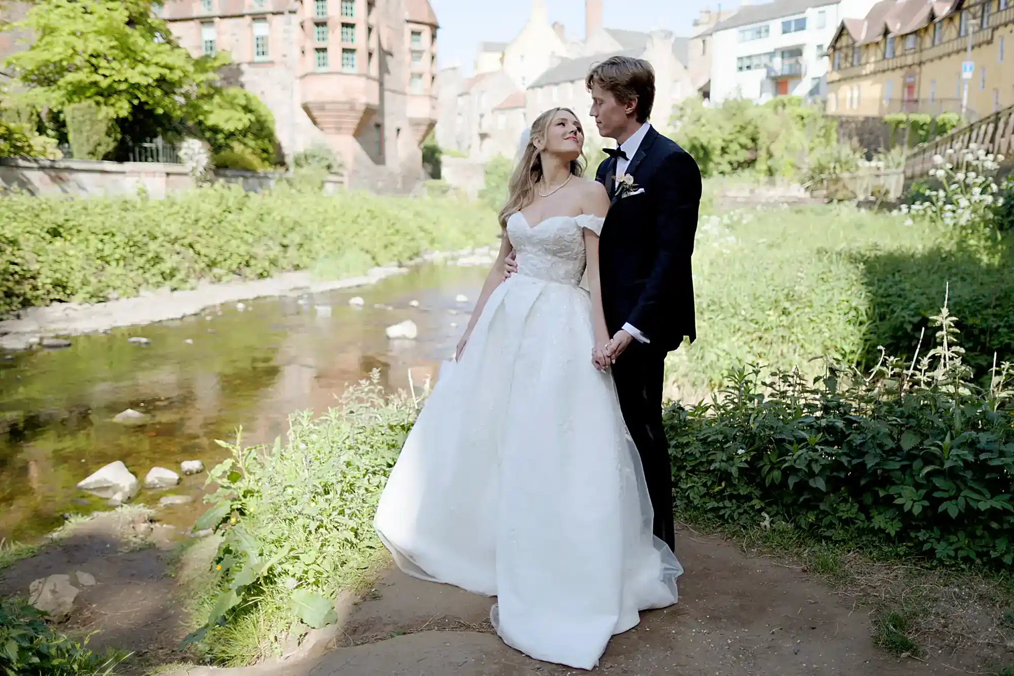 A bride in a white off-the-shoulder gown and groom in a black suit stand holding hands, gazing at each other by a riverside with lush greenery, historic brick buildings, and soft sunlight in the background.