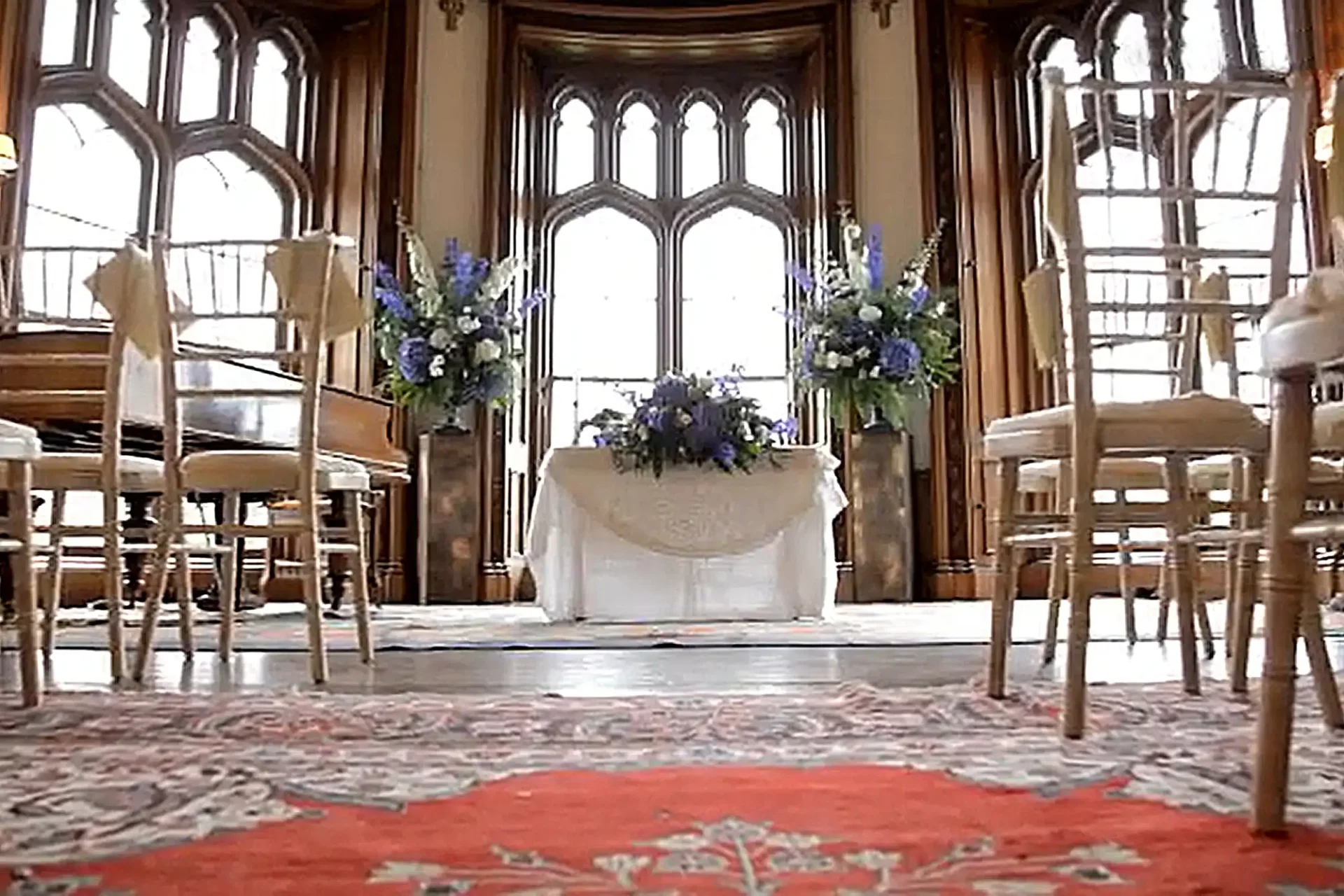 Scottish Borders wedding venues image showing an elegant room at Duns Castle set for a wedding ceremony, featuring a table covered with a white cloth and topped with purple and white flowers, flanked by tall floral arrangements. Wooden chairs face the table, and arched windows let in natural light.