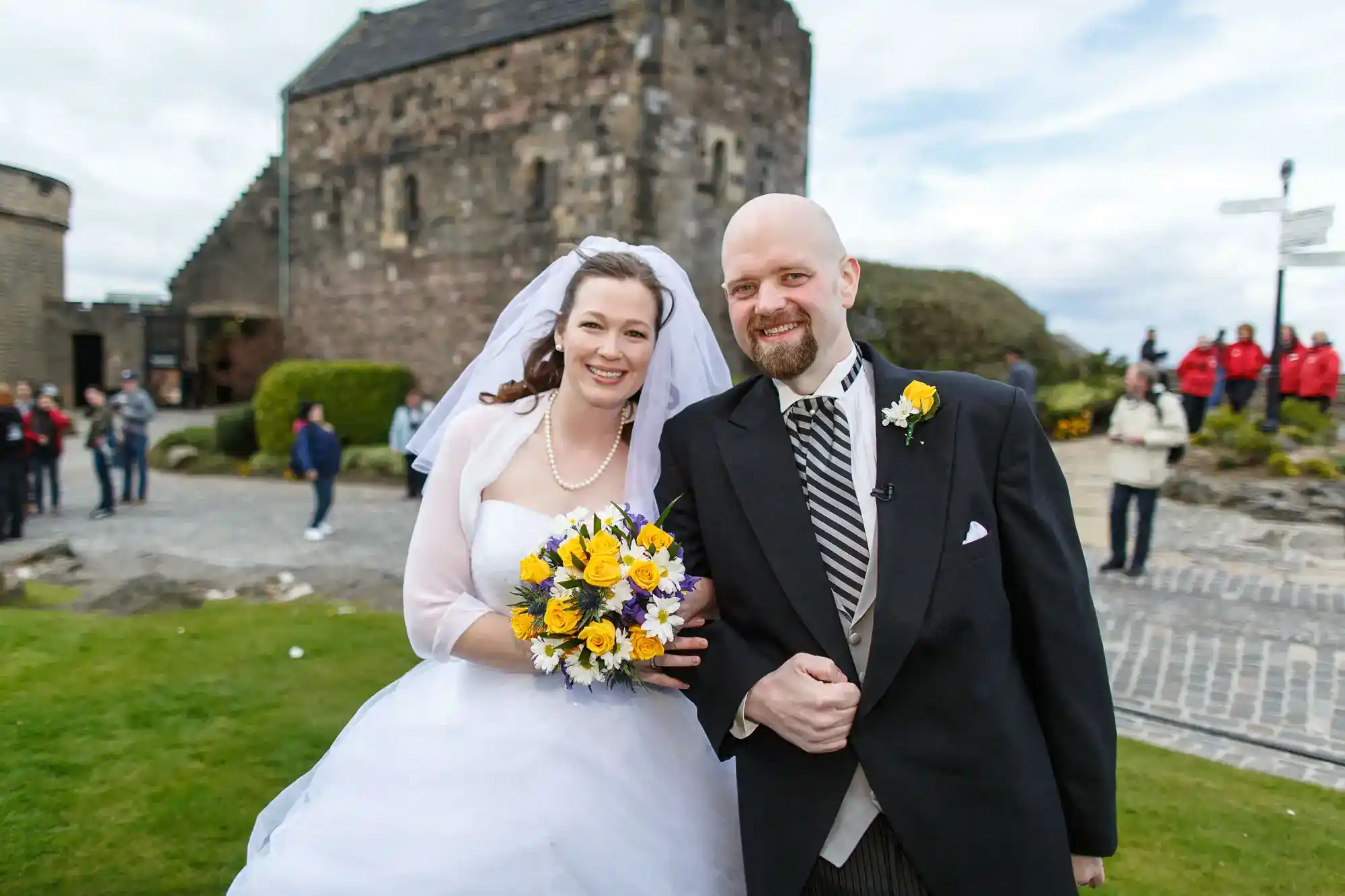 A smiling bride in a white dress and veil holds a yellow and white bouquet, standing arm-in-arm with a bald groom in a black tuxedo outdoors, with an old stone building and people in the background on a cloudy day.