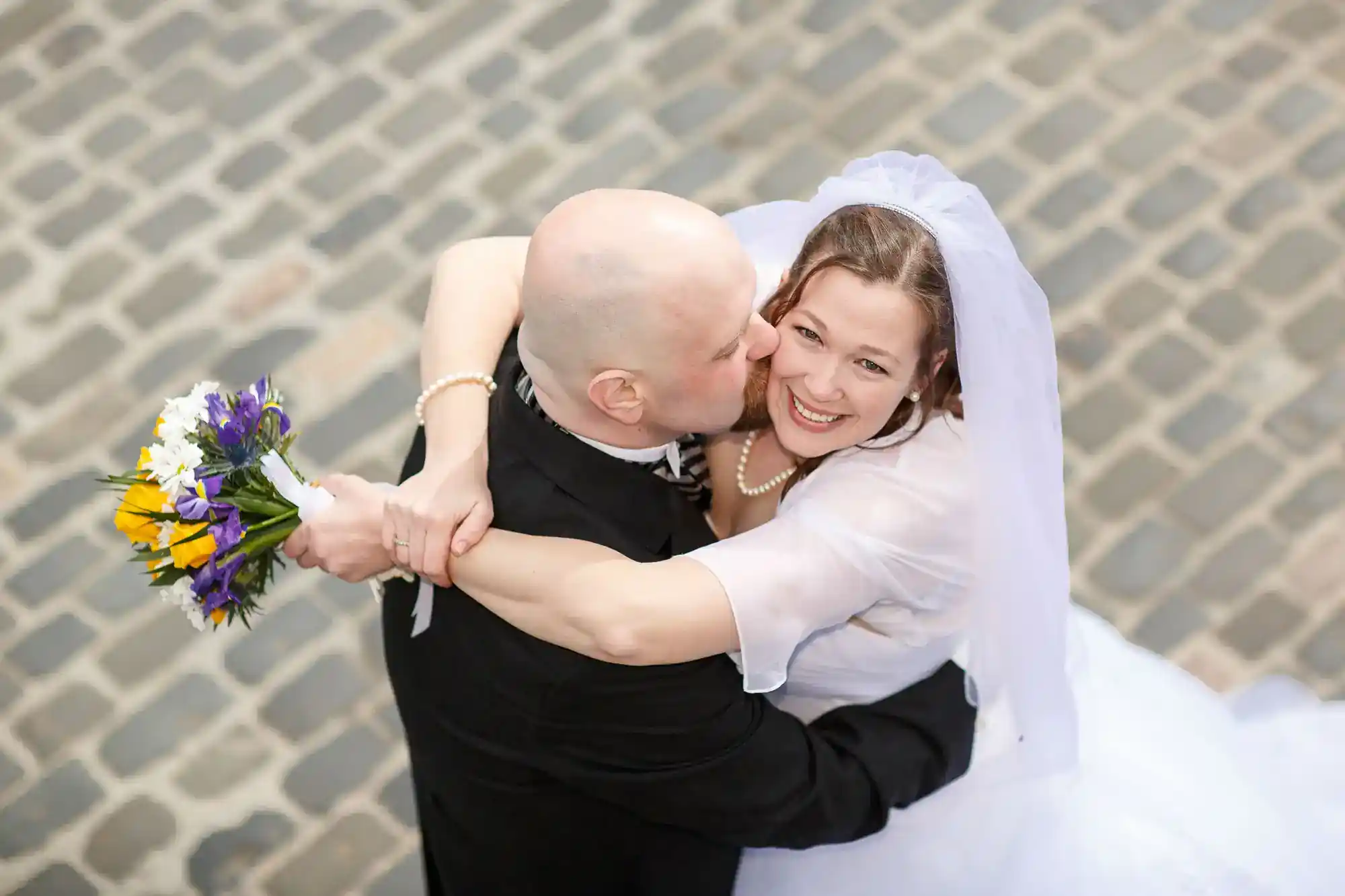A bride in a white dress and veil smiles joyfully whilst being embraced and kissed on the cheek by a bald groom in a black suit on a cobbled street. She holds a bouquet of purple and yellow flowers.