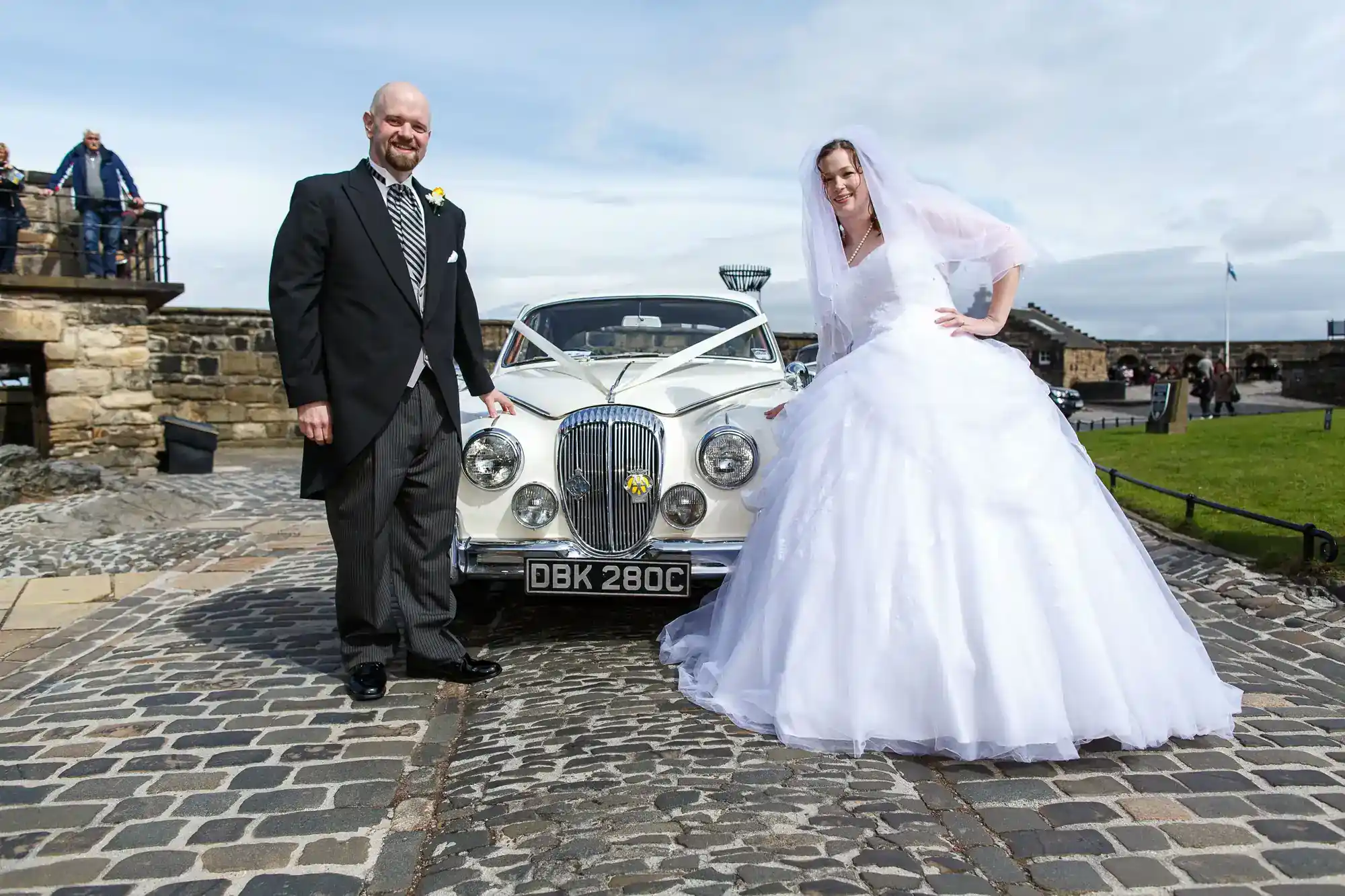 A bride in a white dress and veil stands beside a groom in a black suit and striped tie. They pose by a silver vintage car decorated with white ribbons, on cobbled pavement. Stone buildings and green grass are visible in the background, beneath a cloudy sky.