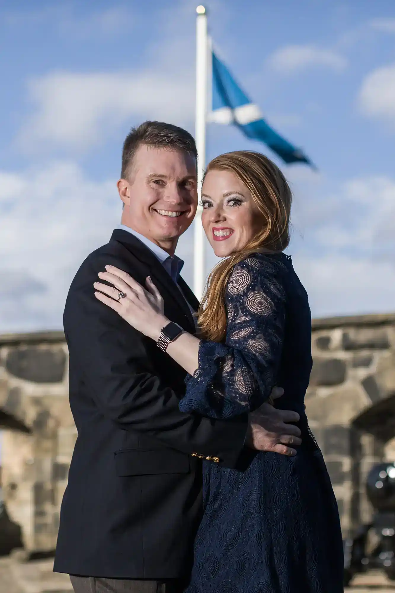 A smiling couple embraces outdoors in front of a stone wall. The woman wears a navy lace dress, and the man wears a dark suit. A flag with a white X on blue flies in the background. The sky is bright with scattered clouds.