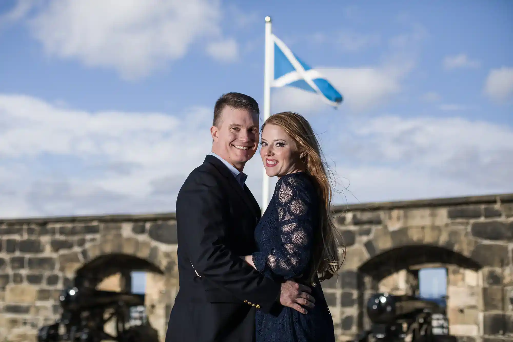 A smiling couple embraces in front of a stone wall with cannon openings. A Scottish flag flies above them against a partly cloudy sky. The man wears a dark jacket, and the woman wears a navy lace dress. Both look at the camera, with the woman’s hair blowing in the wind.