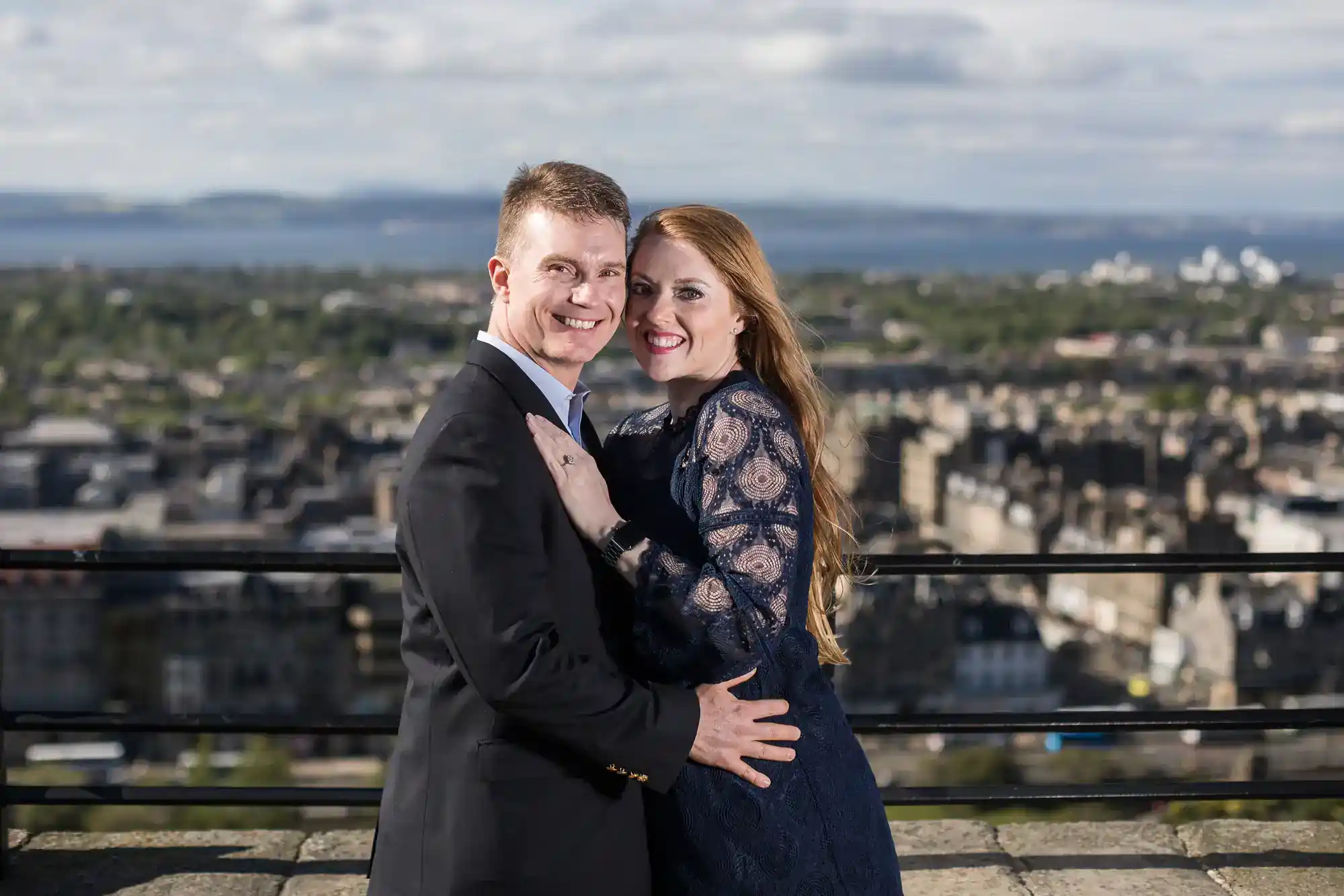 A smiling couple pose together outdoors, dressed formally. The man wears a black suit; the woman wears a navy lace dress. They embrace while standing on a stone terrace, with a cityscape and water in the blurred background under a cloudy sky.