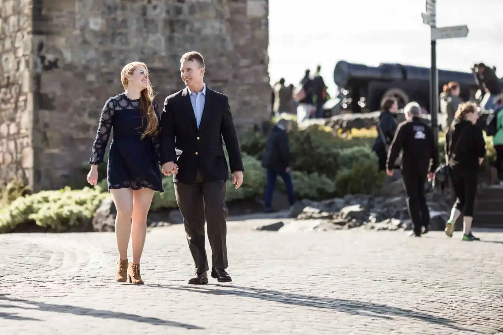 A couple walks hand-in-hand on a cobbled path at St Margaret’s Chapel and Crown Square. The woman wears a dark dress and brown boots; the man wears a suit. In the background, people walk and an old cannon is visible. It’s a sunny day.