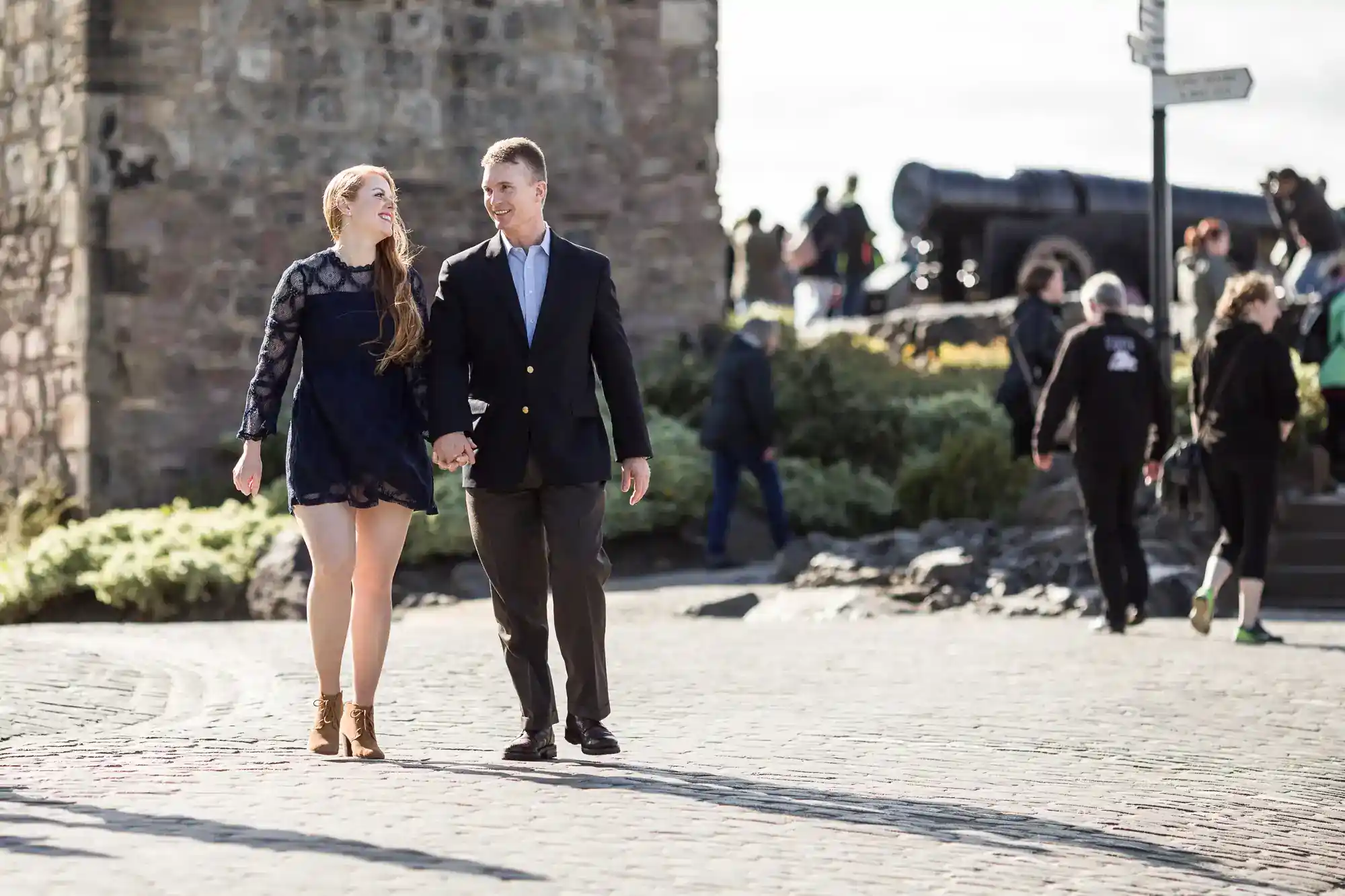 A couple walks hand-in-hand on a cobbled path near a stone wall. The woman wears a dark dress and brown boots; the man wears a suit. In the background, people walk and an old cannon is visible. It’s a sunny day.