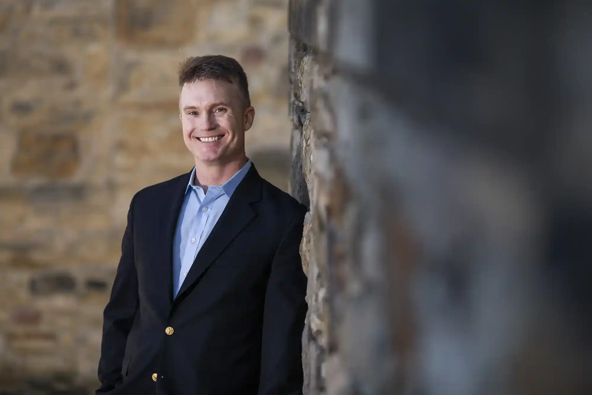 A man in a dark blazer and blue shirt stands smiling against a stone wall. The background is softly blurred, highlighting the textured stones and his casual, confident pose. The lighting is soft, creating a professional yet approachable atmosphere.