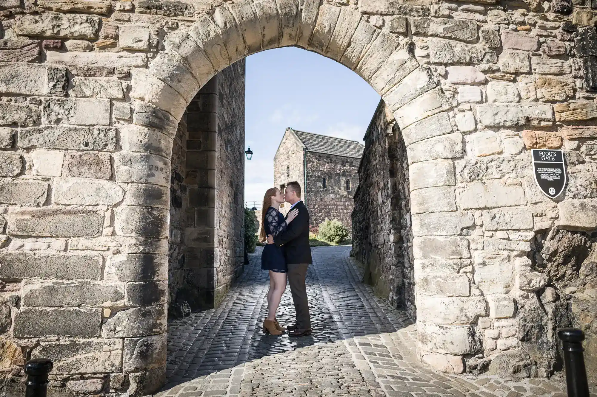 A couple stands on cobblestones under a stone archway at a historic site, kissing and embracing. Sunlight highlights the scene, with old stone buildings and a blue sky in the background. A sign is visible on the right side of the archway.