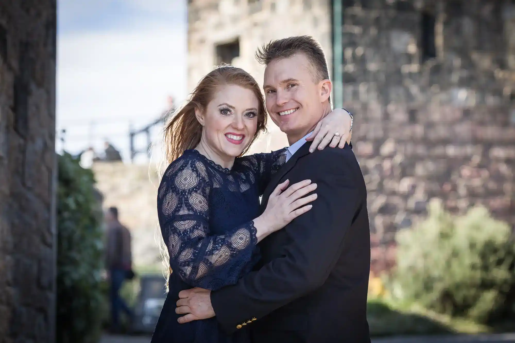 A smiling couple pose closely outdoors by a stone building. The woman, with long red hair in a navy lace dress, wraps her arm around the man’s shoulder. The man, in a dark suit, hugs her waist. They both look at the camera, appearing happy and affectionate.