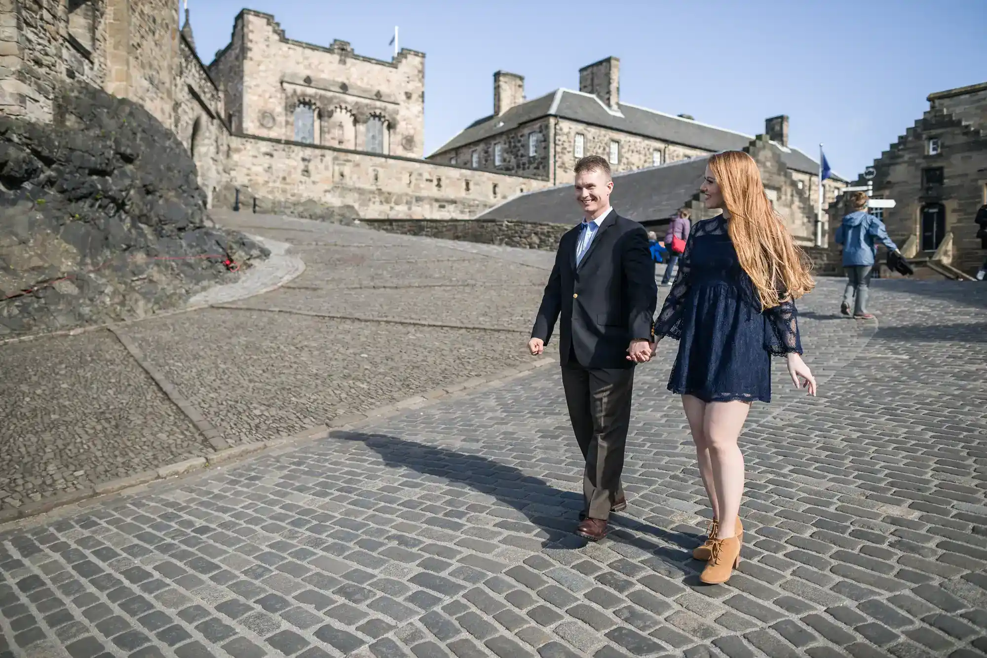 A smiling couple walk hand-in-hand on a cobbled path near an old stone castle. The man wears a dark suit; the woman wears a navy dress and tan boots. Other people are seen in the background under a clear blue sky.
