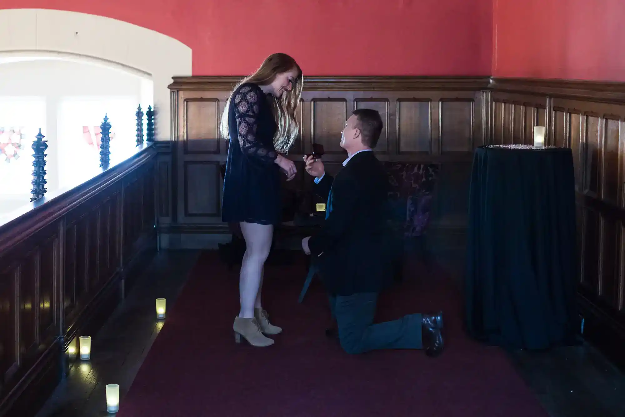 A man in a suit kneels on one knee, proposing to a woman in a black dress and tan boots. They are indoors, near a window, with candles on the floor and a small table covered in black cloth nearby. The mood is romantic and intimate.