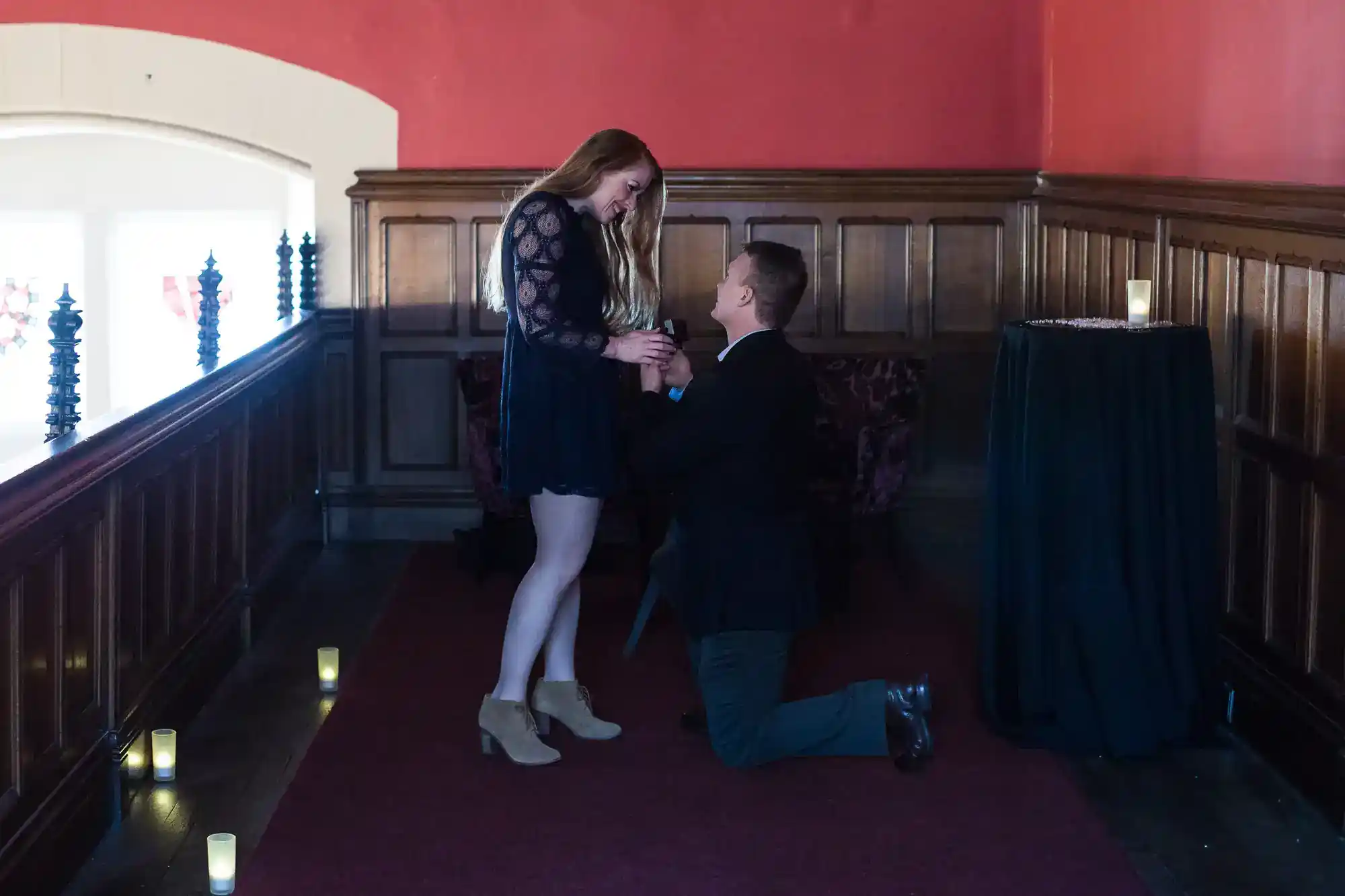 A man kneels and holds a woman’s hands in a proposal. She smiles, wearing a black dress and boots. The setting is an elegant, dimly lit room with red walls, wood panelling, and candles lining the floor.