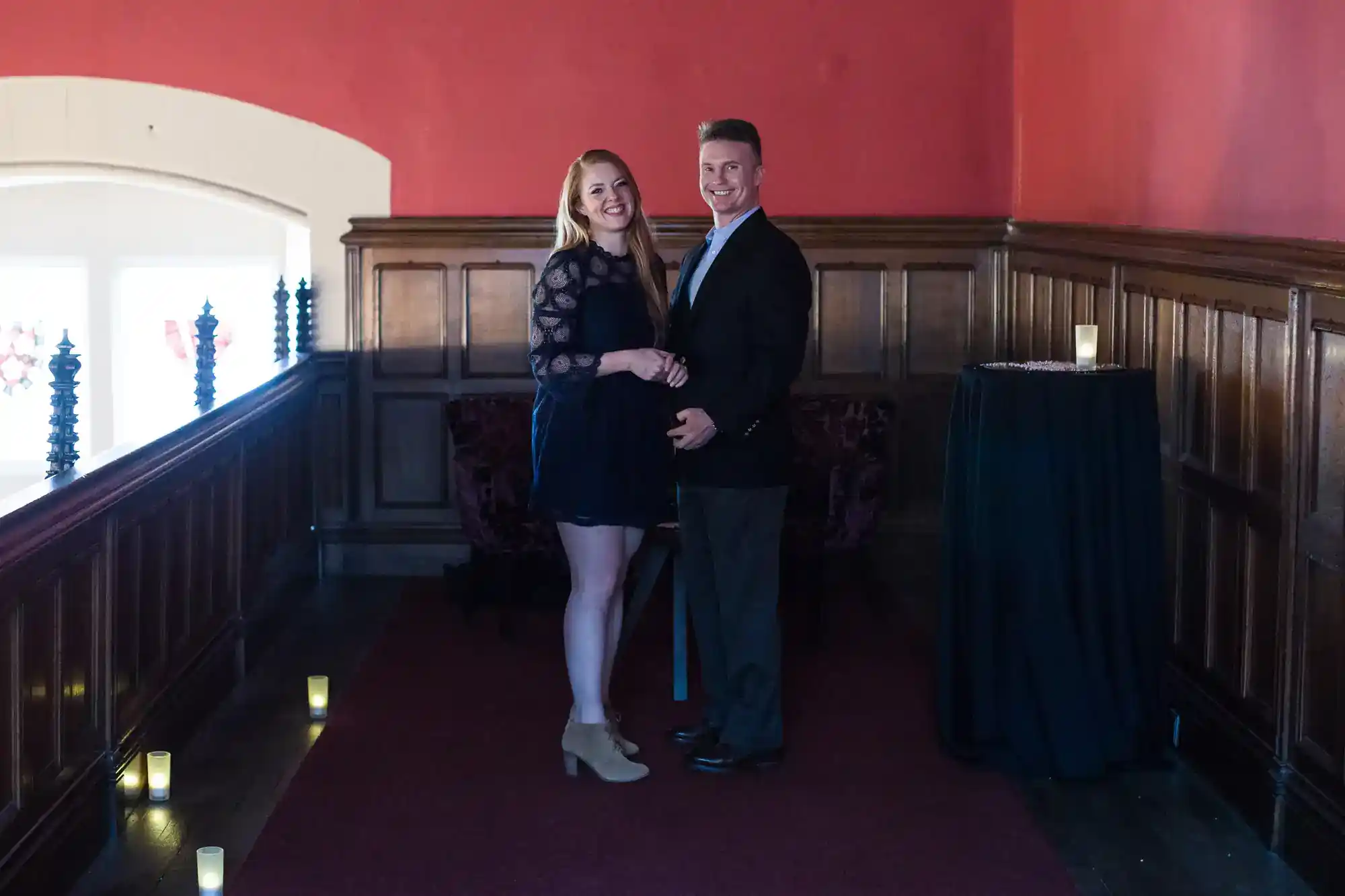 A smiling couple stands in a carpeted corridor with red walls and dark wood panelling. The woman wears a black lace dress and tan shoes; the man wears a suit. Small candles line the floor, and a tall round table draped in black is on their right.