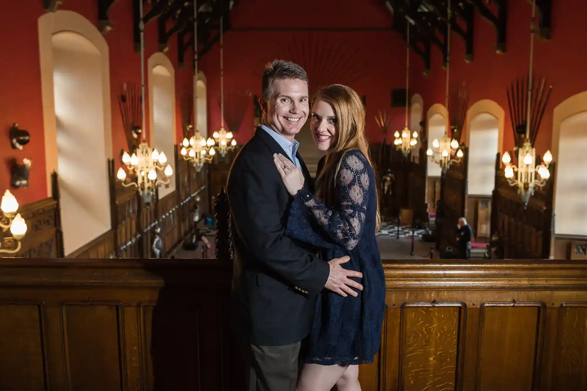 A smiling couple pose closely together on a balcony in a grand hall with red walls, chandeliers, arched windows, and dark wooden panelling. The man wears a suit and the woman wears a navy lace dress. The background shows people seated below.