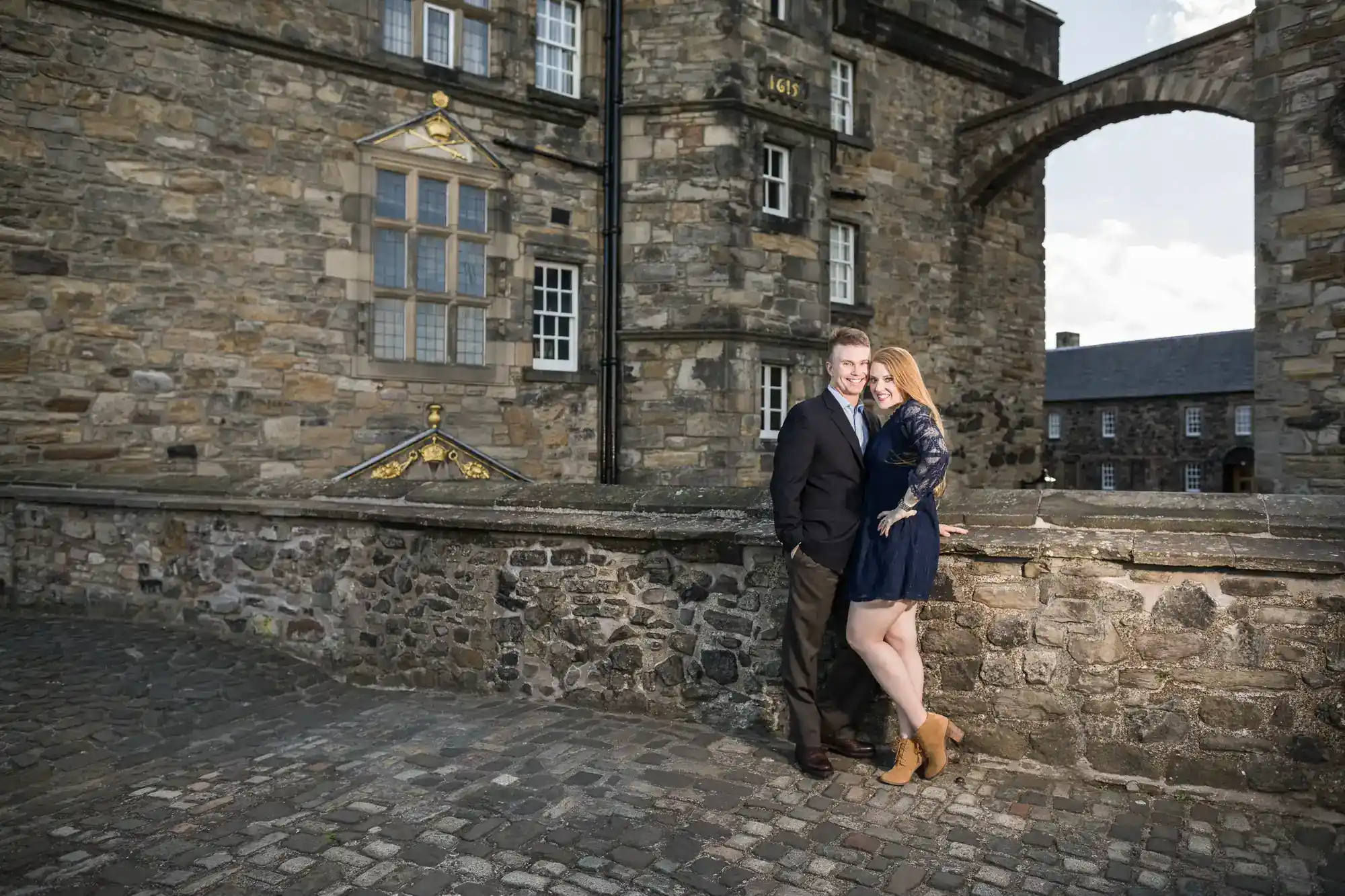 A couple stand close together, smiling, by a stone wall in front of a historic stone building with many windows. The man wears a suit; the woman wears a navy dress and tan boots. The setting appears to be an old European castle or fortress.