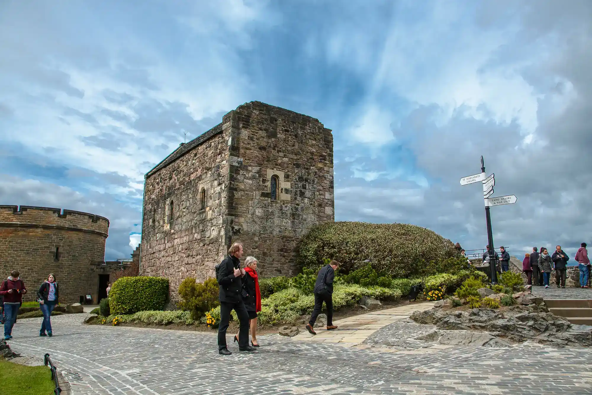 Visitors walk near a historic stone building with small arched windows, surrounded by trimmed bushes and a cobbled path. A signpost stands nearby. The sky is partly cloudy, and several people are seen exploring the area.