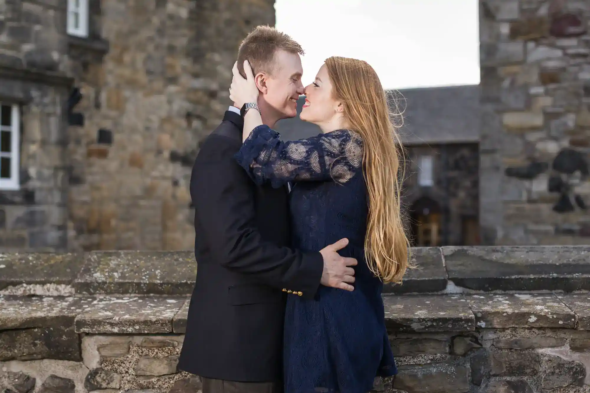 A couple stands close in front of a stone building. The woman, in a dark blue dress, holds the man's face and smiles up at him. The man, in a dark suit, embraces her round the waist. Both appear happy and affectionate outdoors.